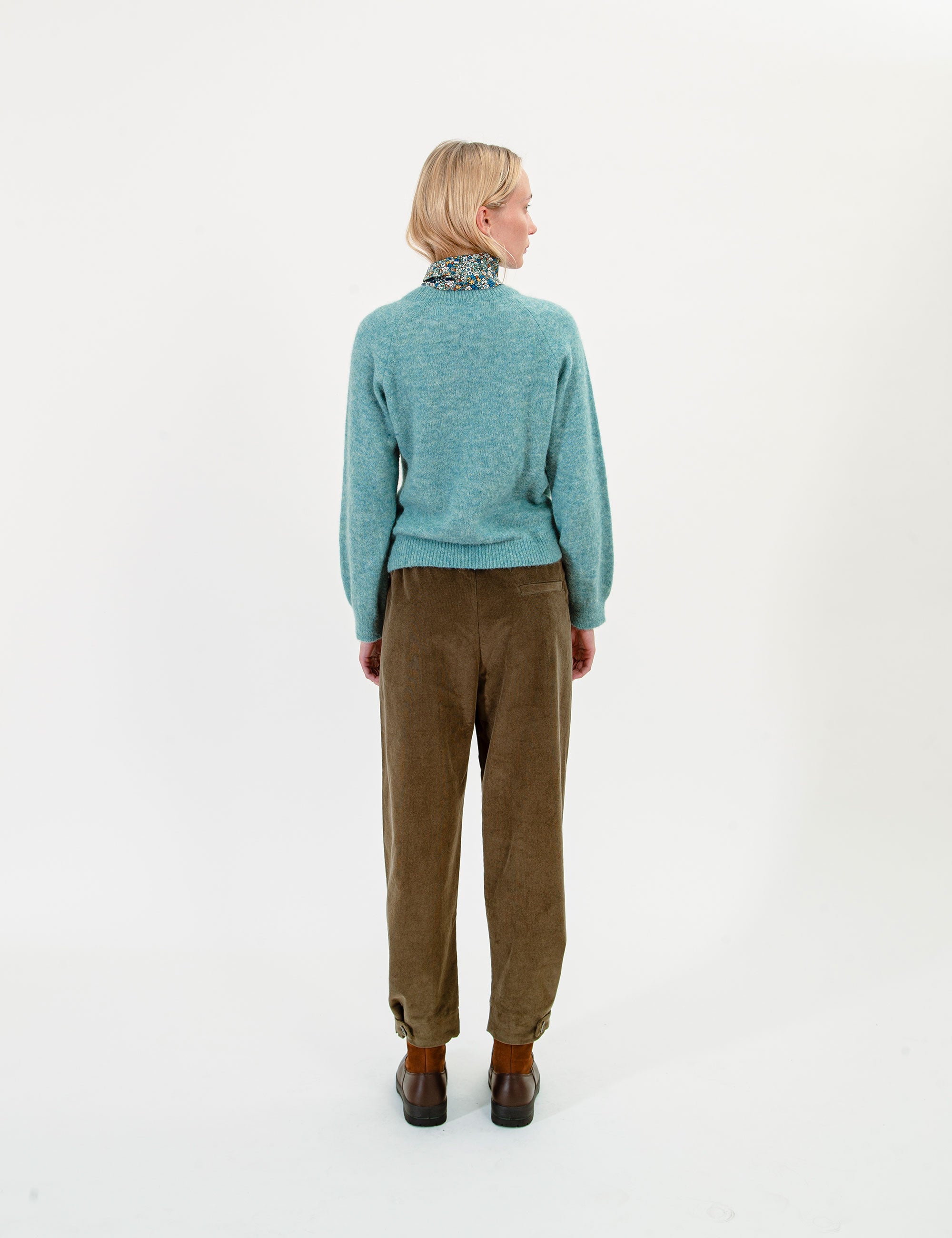 Woman wearing a teal sweater and olive green corduroy pants with a patterned scarf on a white background