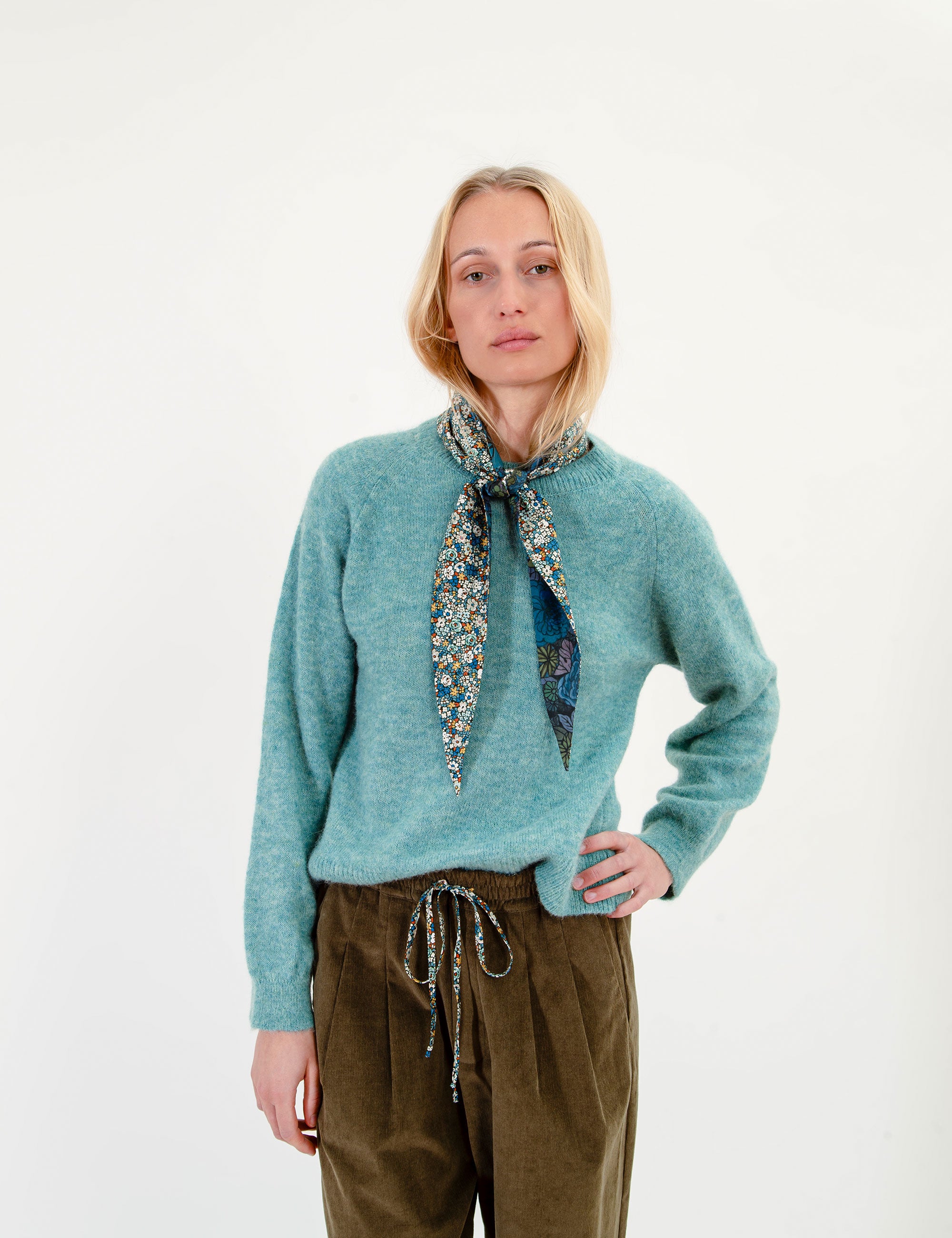 Woman wearing a teal sweater and olive green corduroy pants with a patterned scarf on a white background
