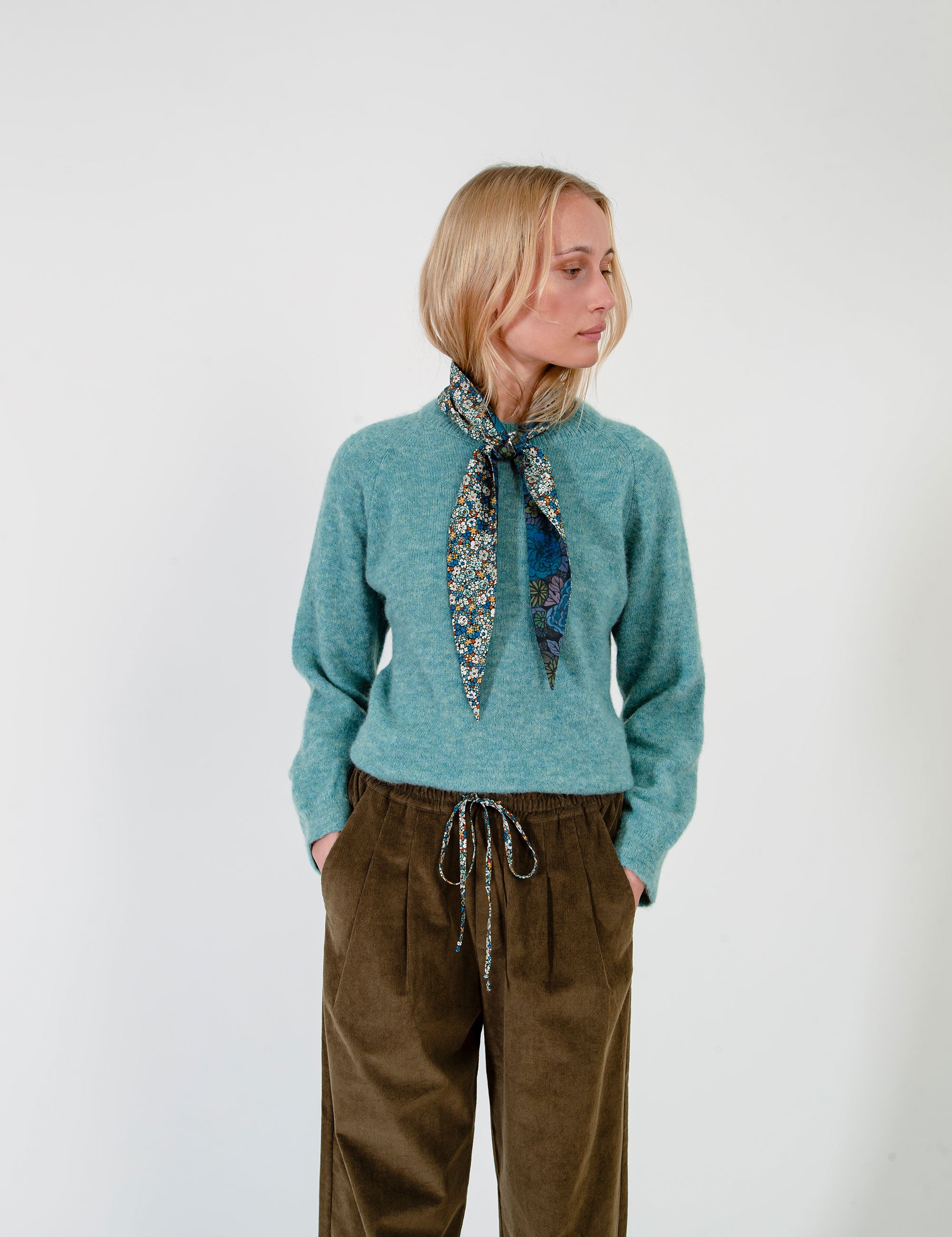 Woman wearing a teal sweater, olive green corduroy pants, and a patterned scarf on a white background
