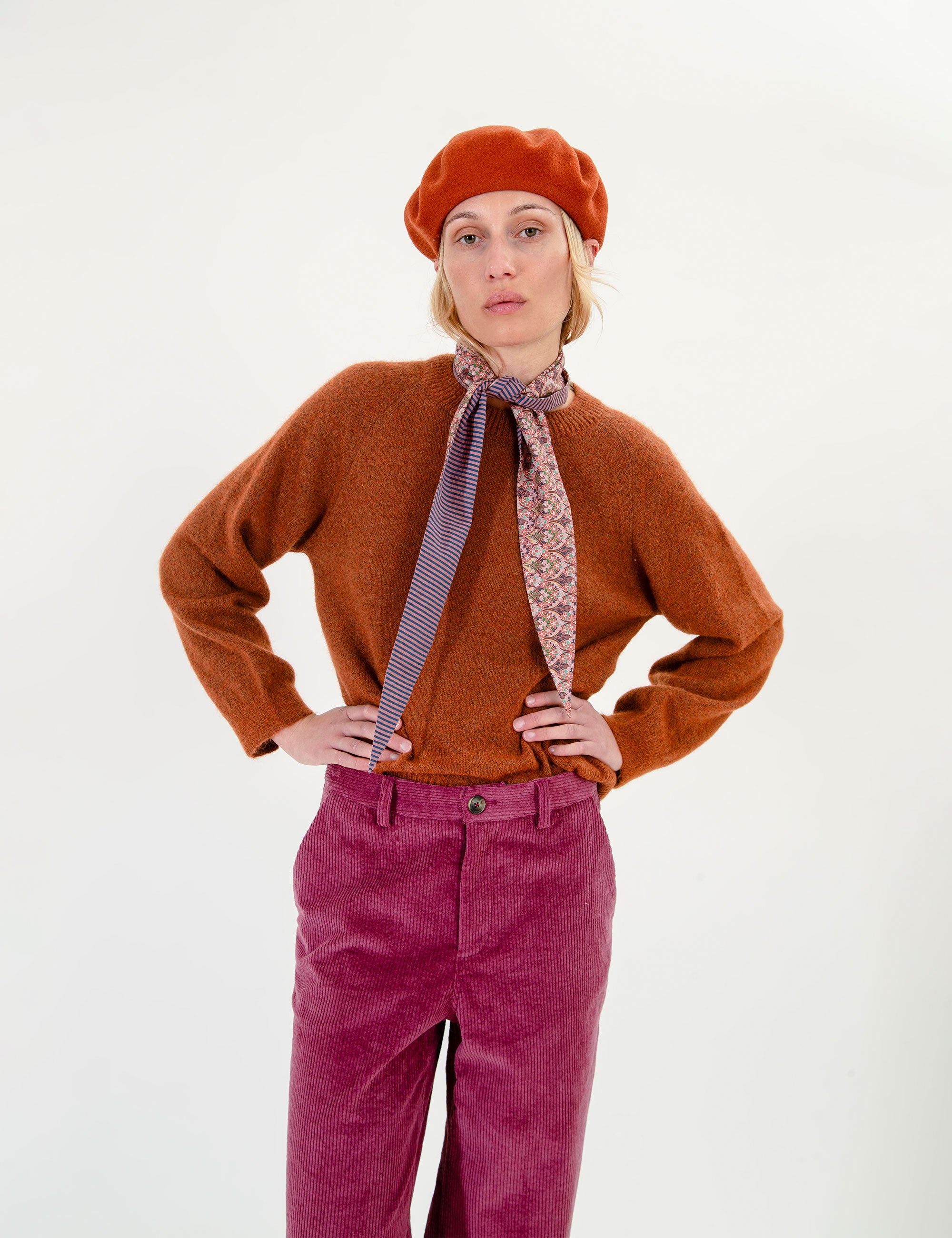 Woman wearing a brown sweater, red beret, and pink pants with a patterned scarf on a white background