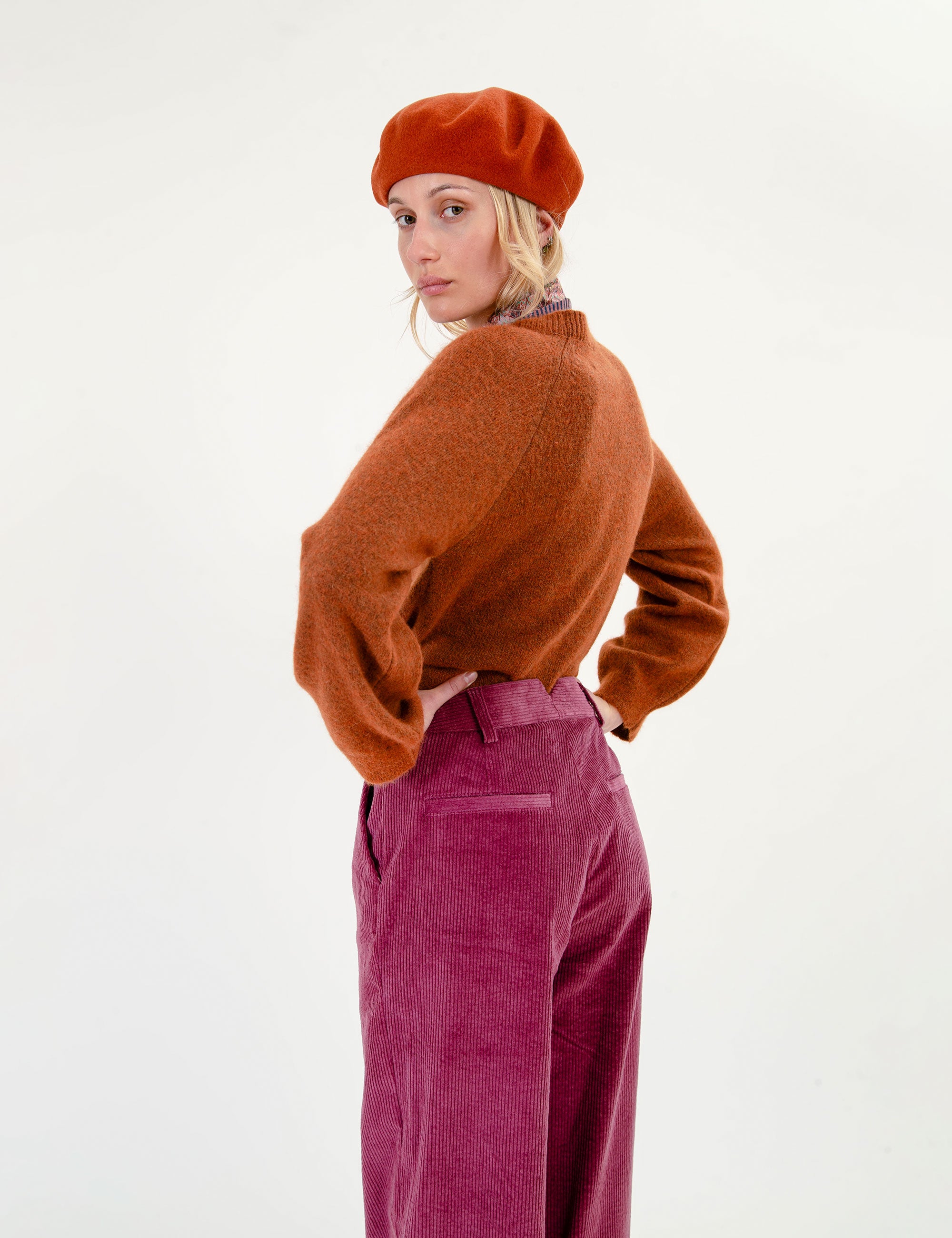 Woman wearing a brown sweater, red beret, and pink corduroy pants on a white background