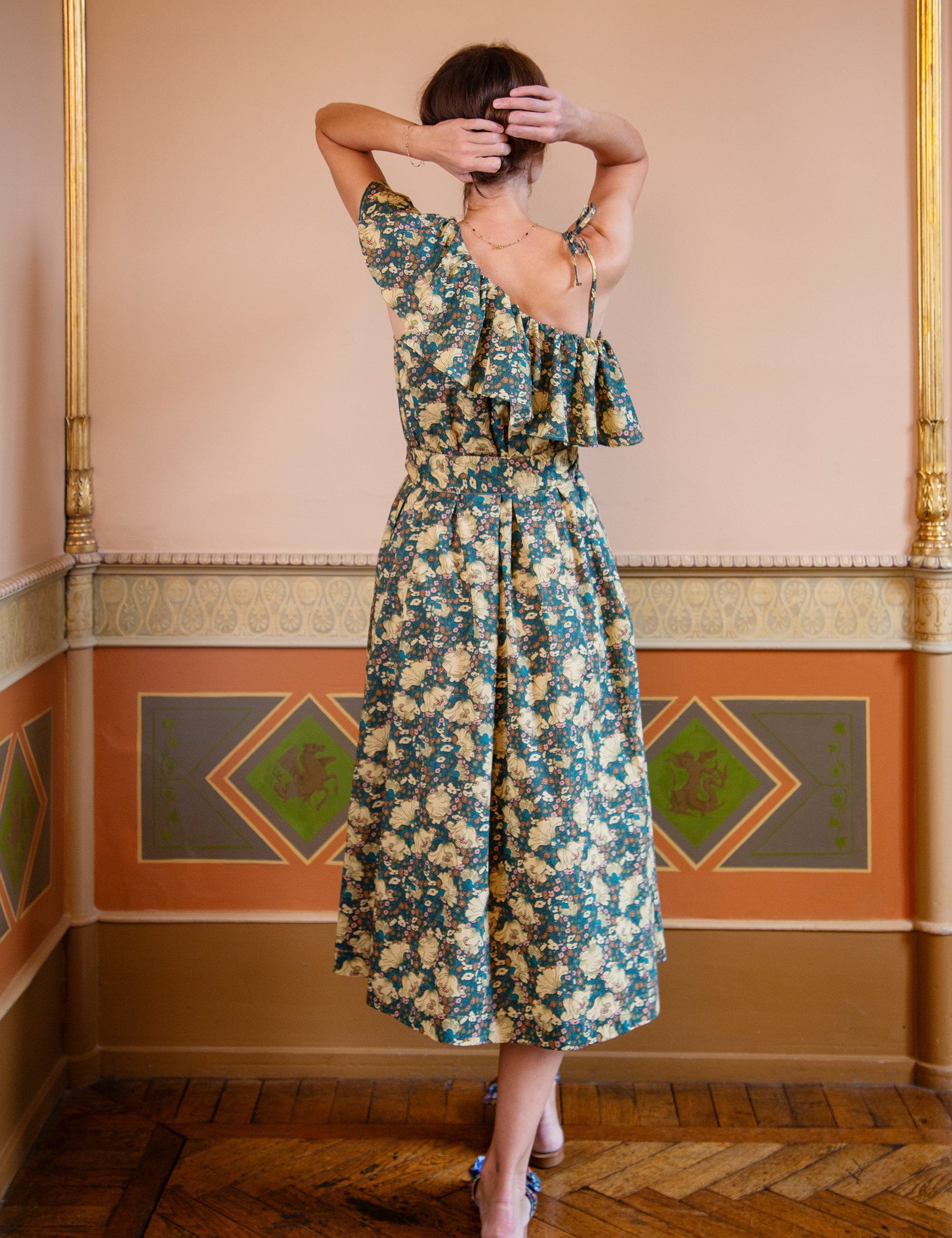 Woman wearing a green floral patterned one shoulder dress in an ornate room with decorative walls.