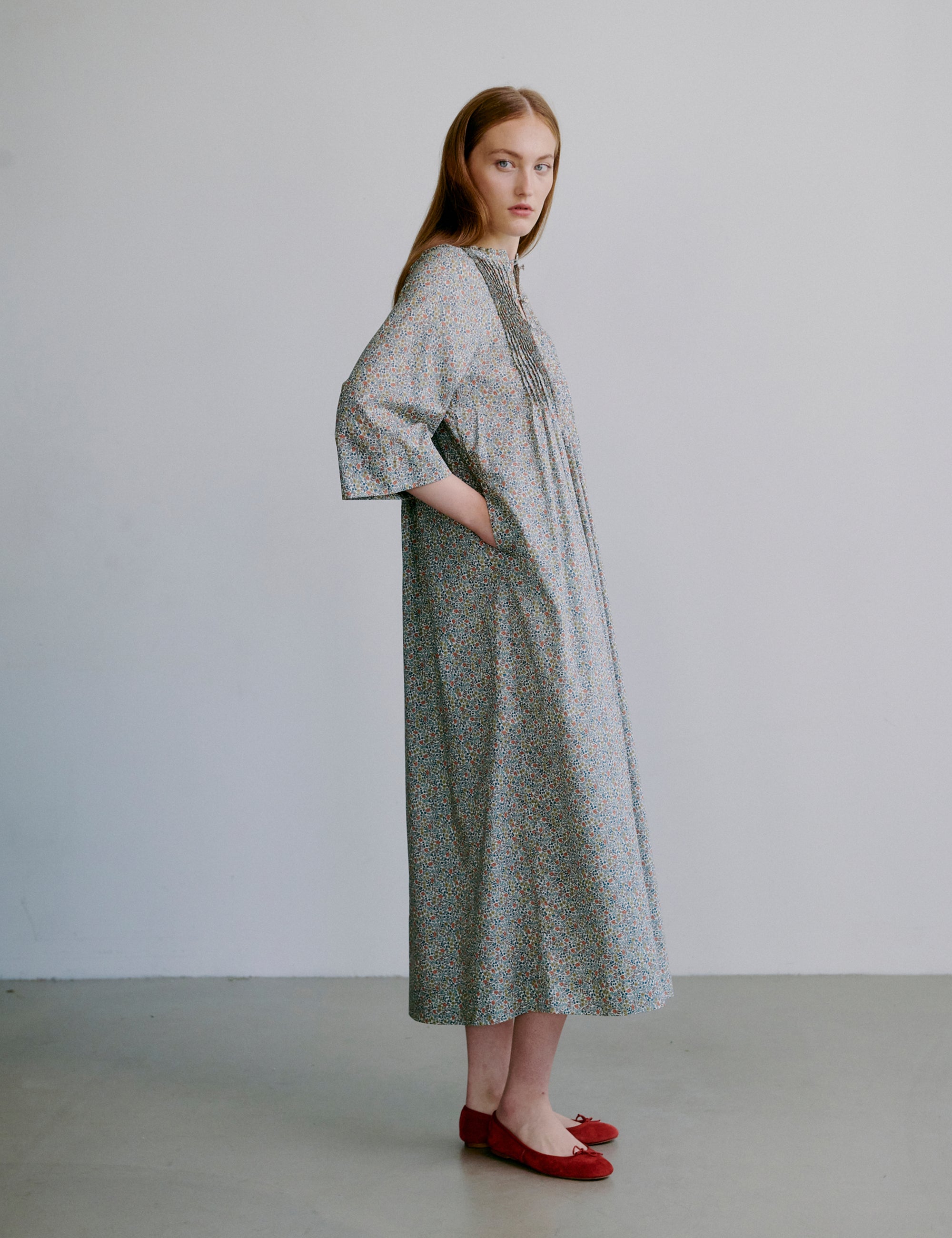 Woman wearing a long, light-colored dress with a subtle floral pattern, semi-long sleeves and red shoes