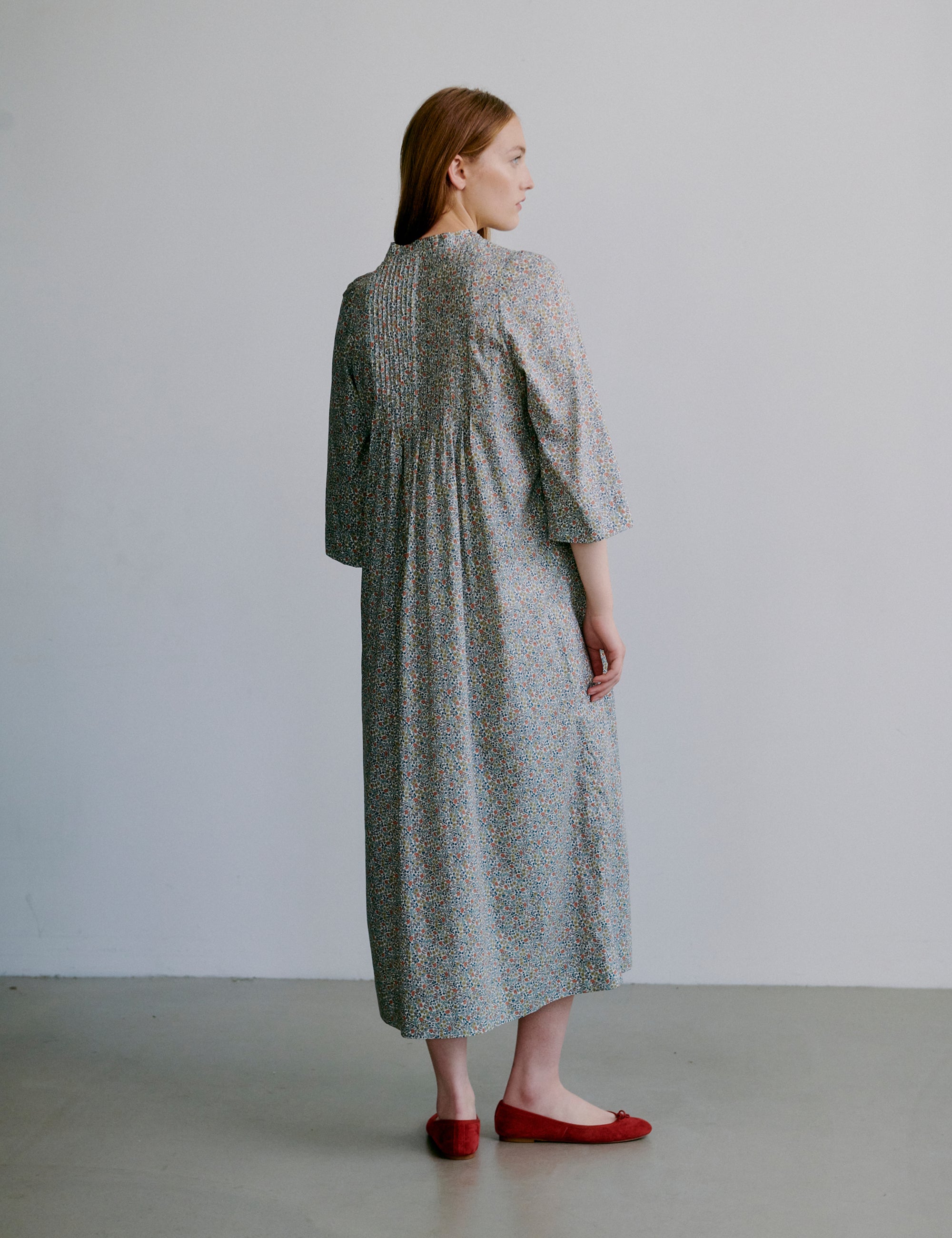 Woman wearing a long, light-colored dress with a subtle floral pattern, semi-long sleeves and red shoes