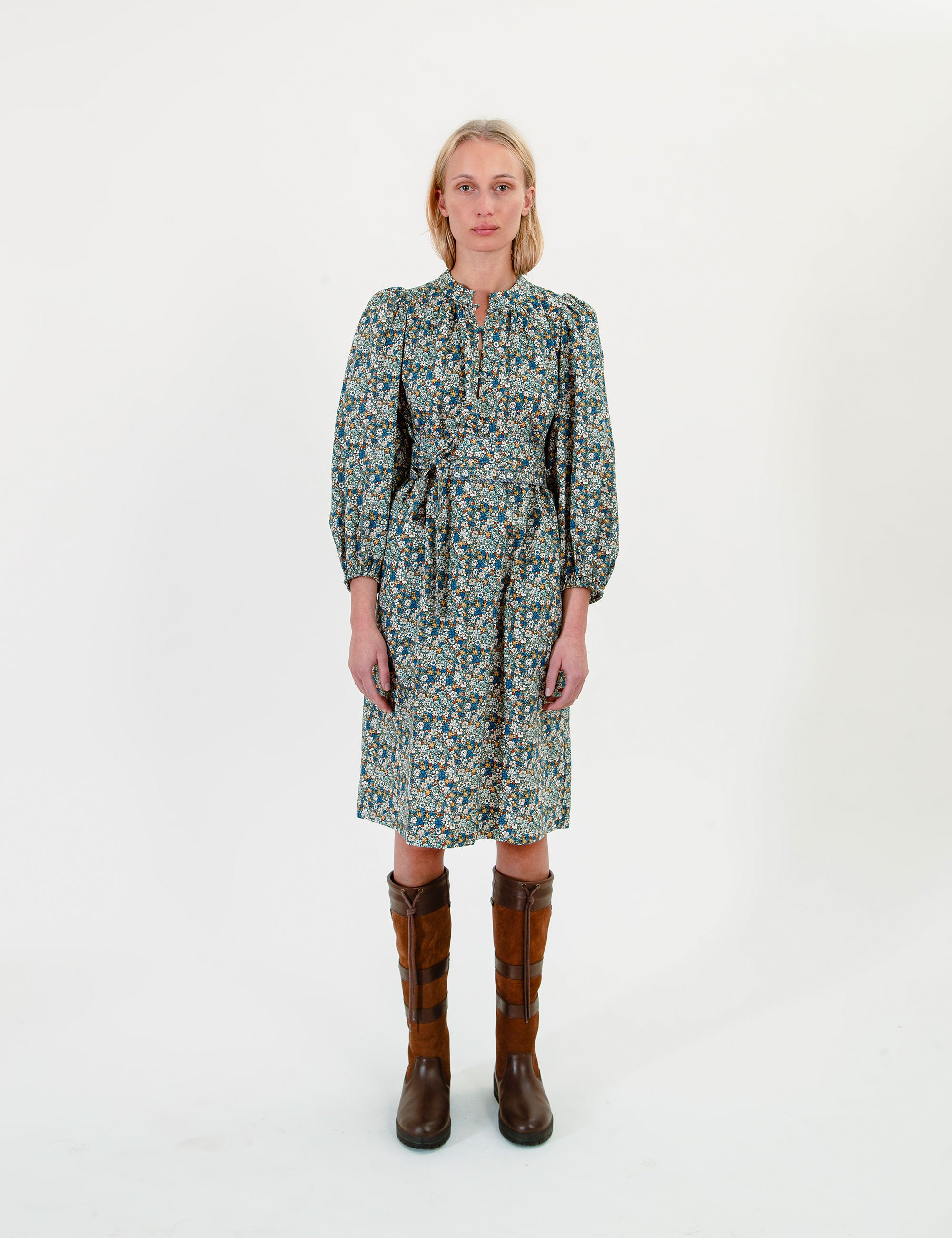 Woman wearing a short green-blue floral dress with long sleeves and a belt on a white background