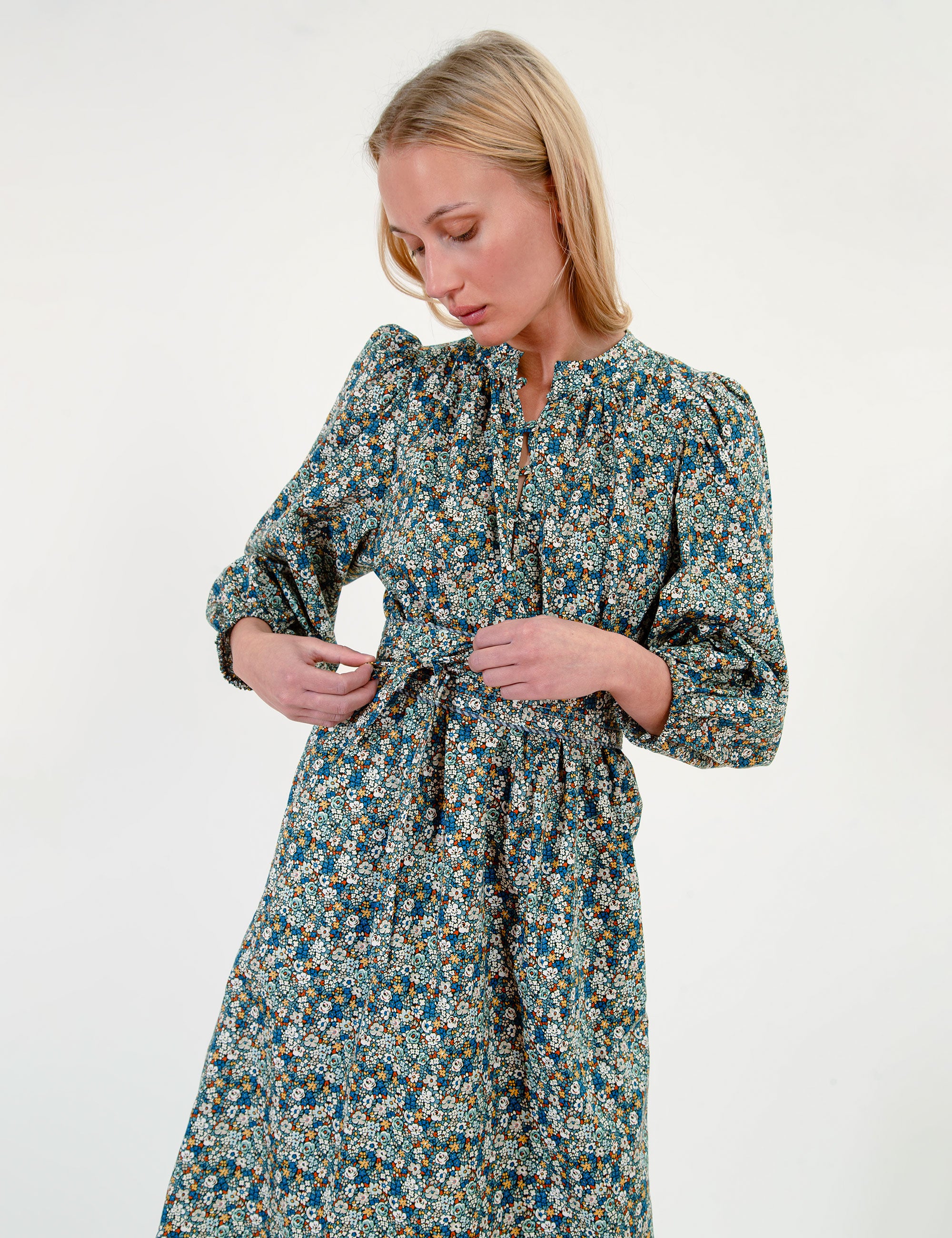 Woman wearing a short green-blue floral dress with long sleeves and a belt on a white background