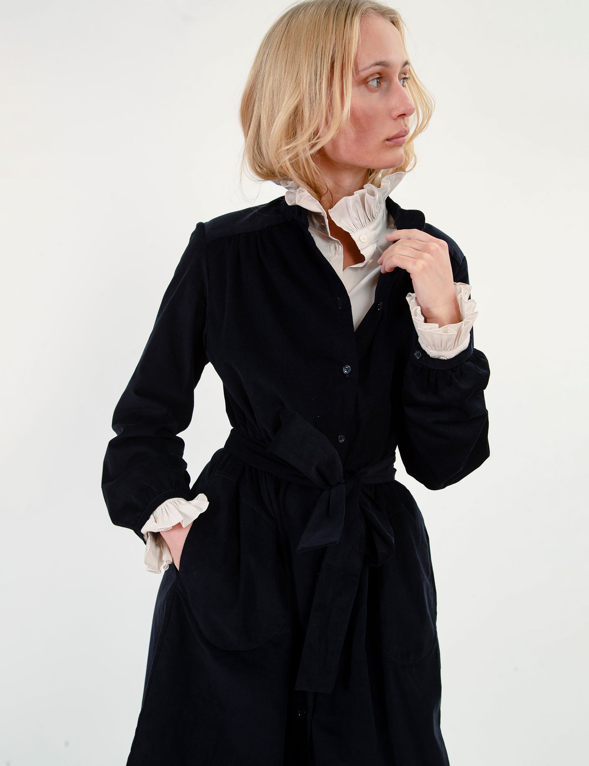 Woman wearing a blue corduroy dress  over a white shirt with a frilled high collar on a white background