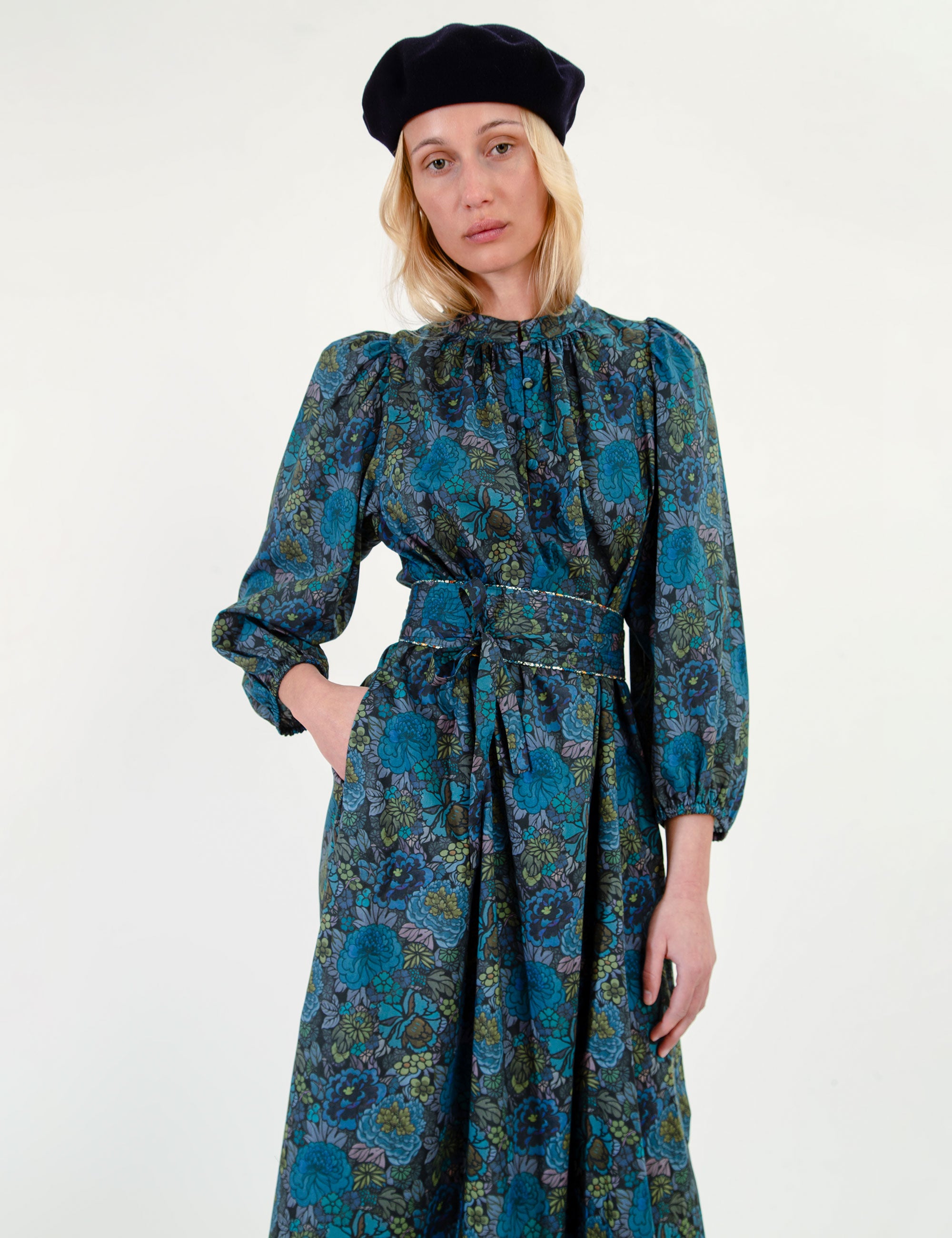 Woman wearing a blue and green floral dress with long sleeves and a belt, standing against a white background.