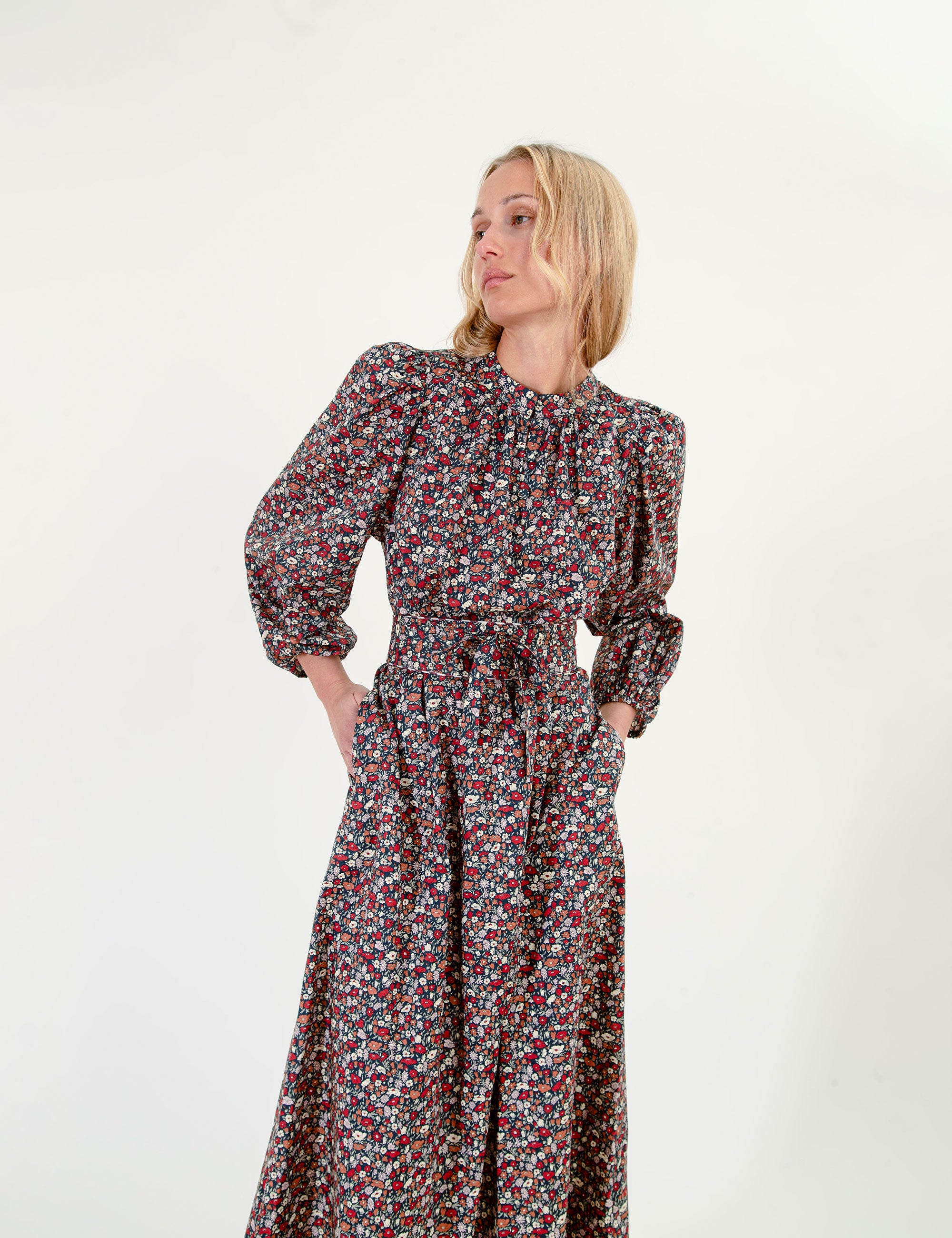 A woman modeling a long-sleeved midi dress with a dense small floral print in red, green, and white on a dark background, set against a plain white studio backdrop.