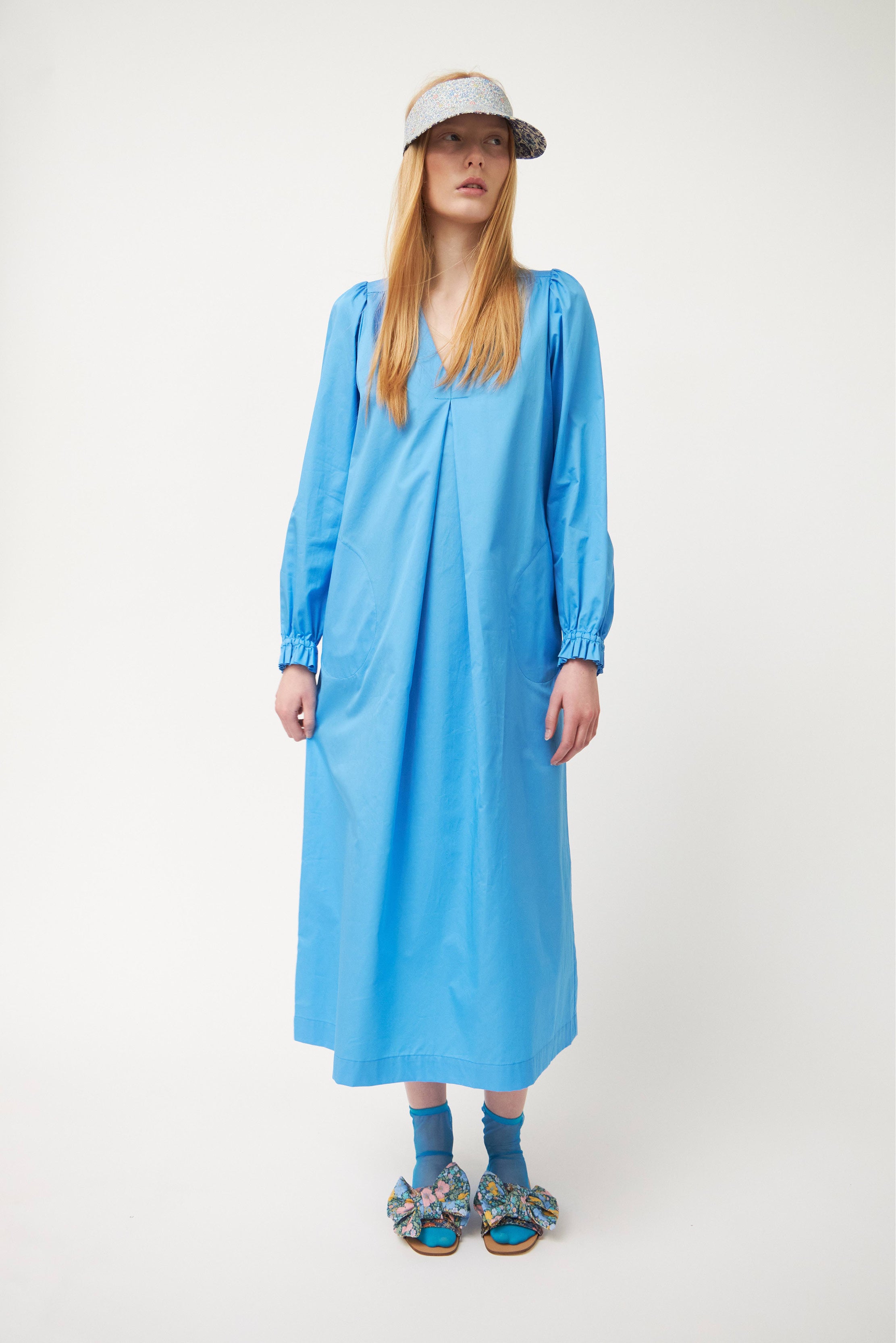 Woman in long blue cotton tunic dress with v-neck, puffed long sleeves and ruffled cuffs with sun shade cap and sandals with bows