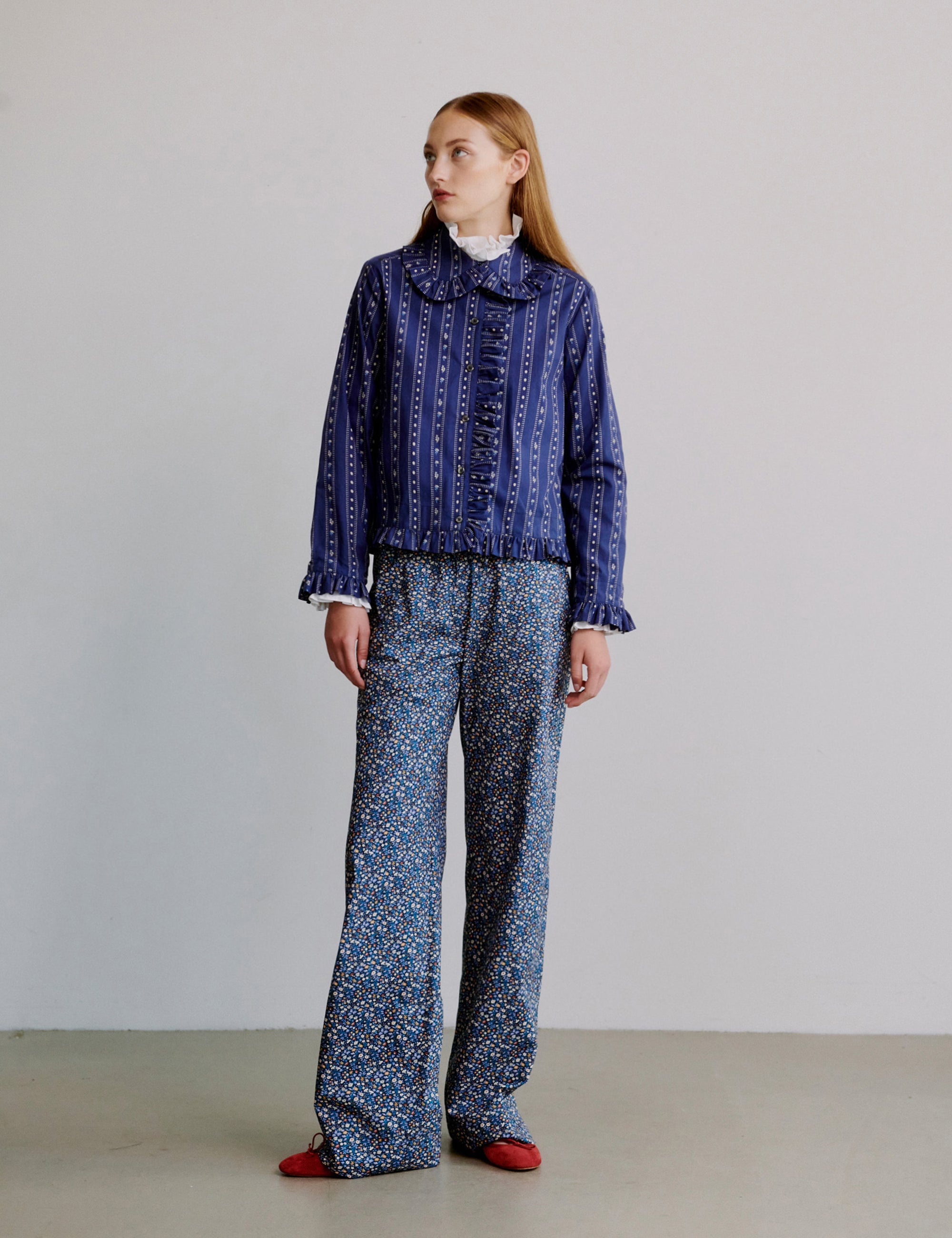 Woman wearing a blue patterned shirt with frill collar and floral patterned blue pants on a plain background