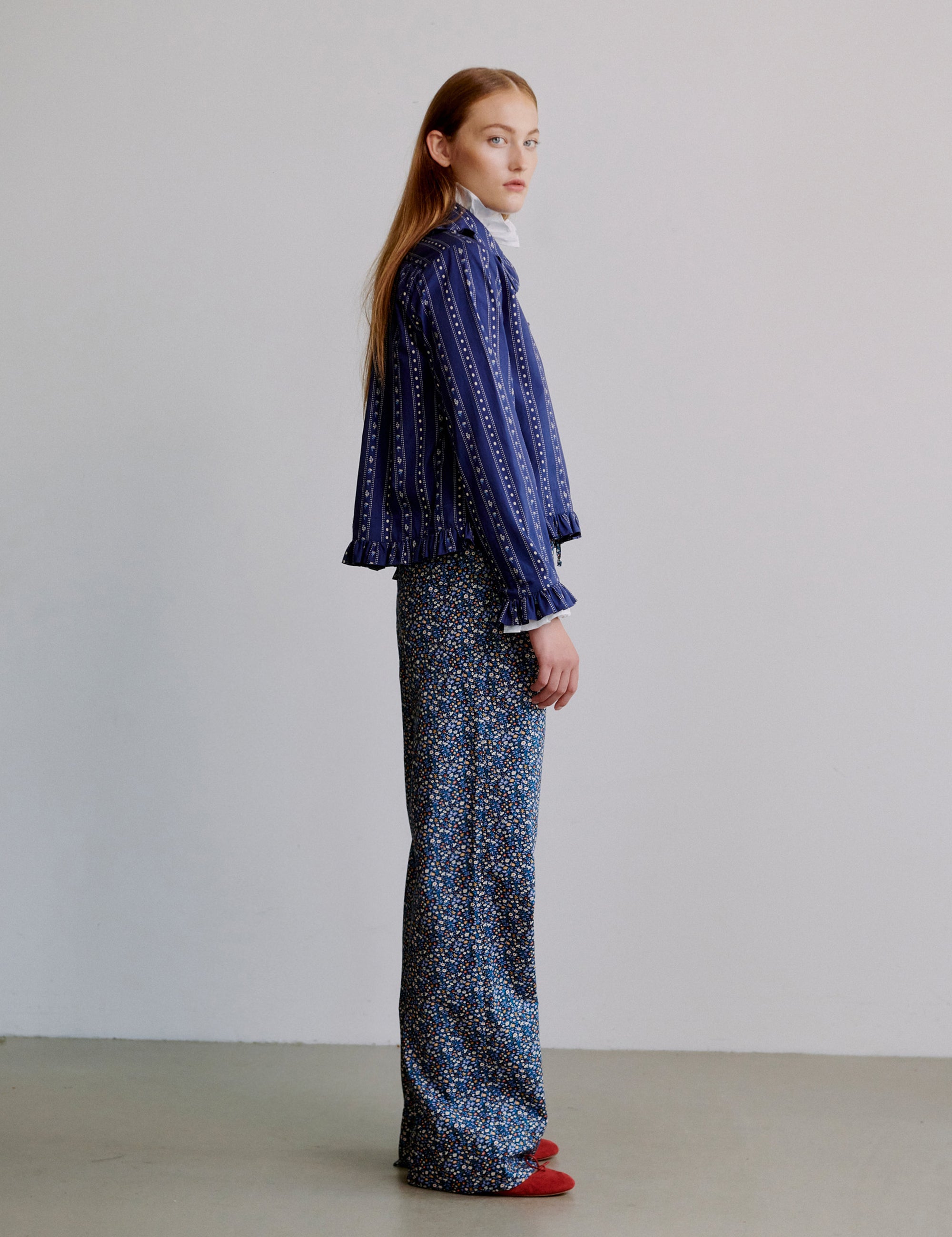 Woman wearing a blue patterned shirt with frill collar and floral patterned blue pants on a plain background