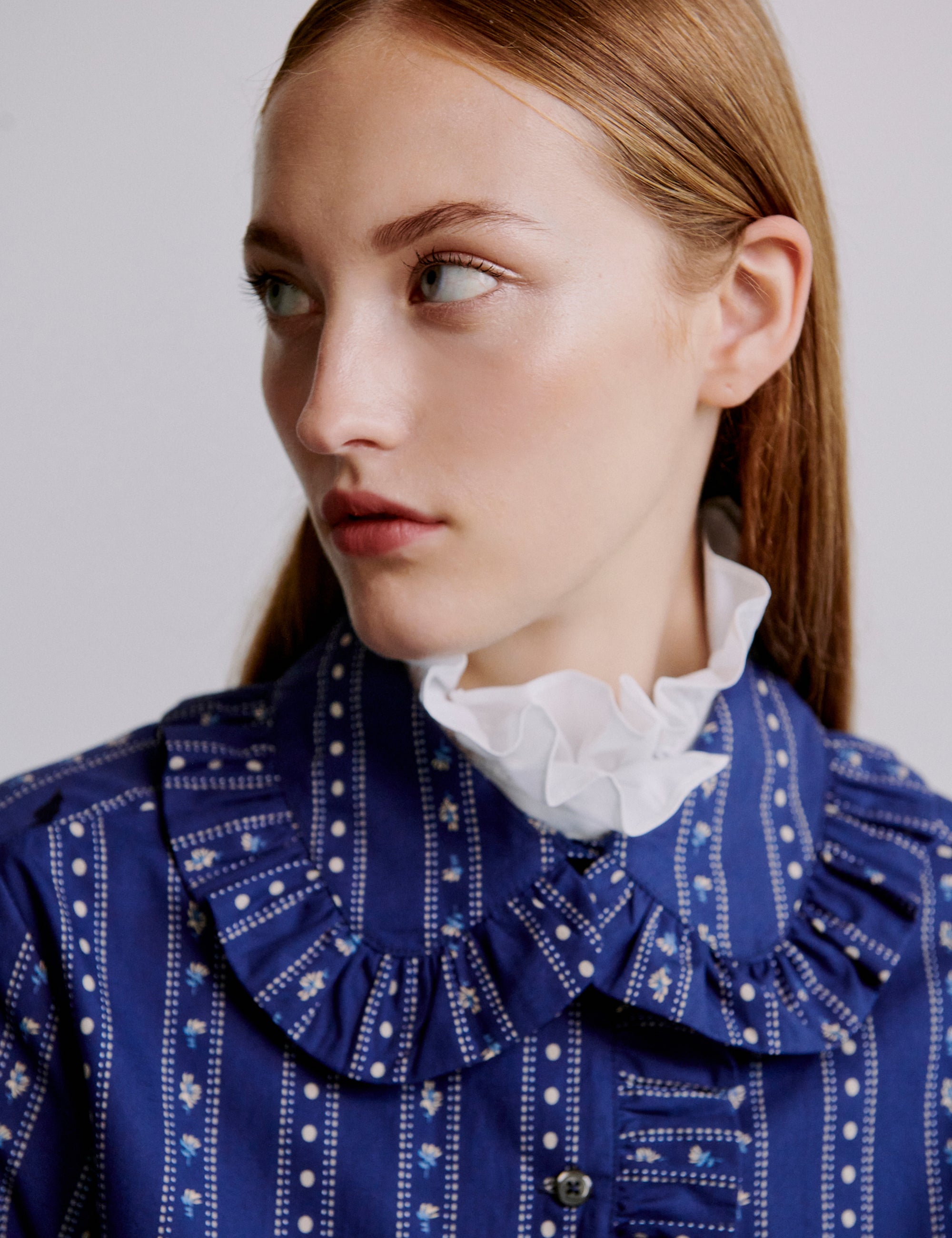 Woman wearing a blue patterned jacket with frilled collar over a white collar against a plain background