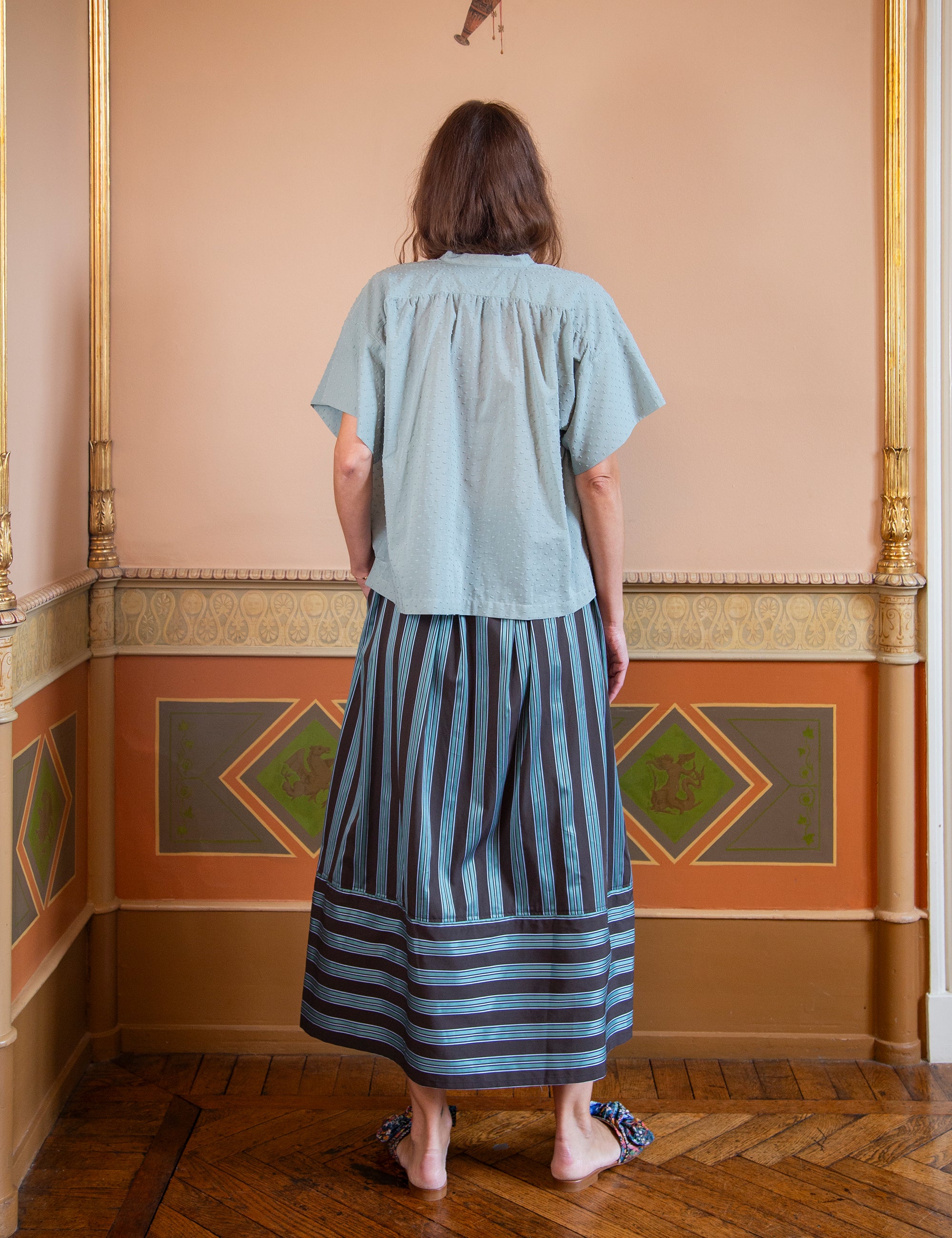 Woman wearing a light green top and striped maxi skirt standing in an ornate room.