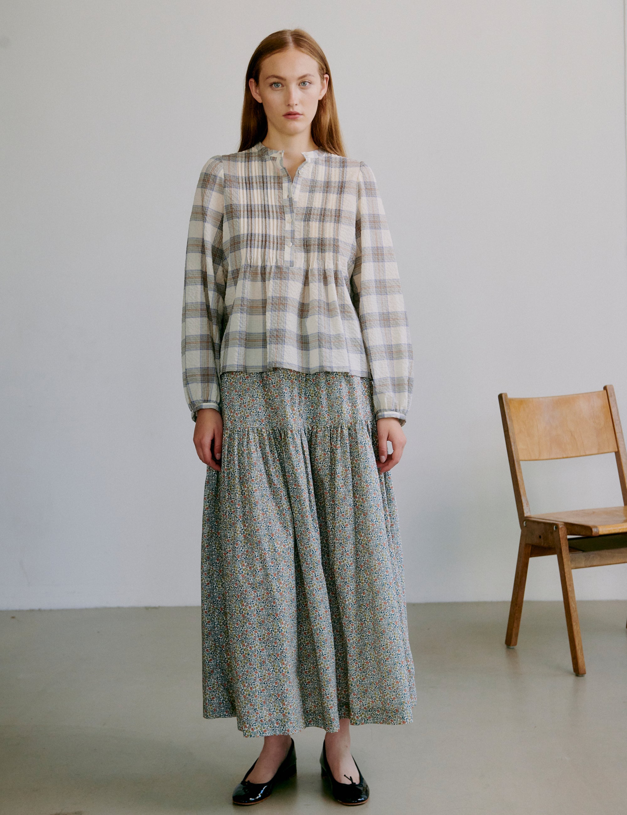 Woman wearing a check patterned white and grey shirt and long floral skirt standing in a minimalistic room with a wooden chair.