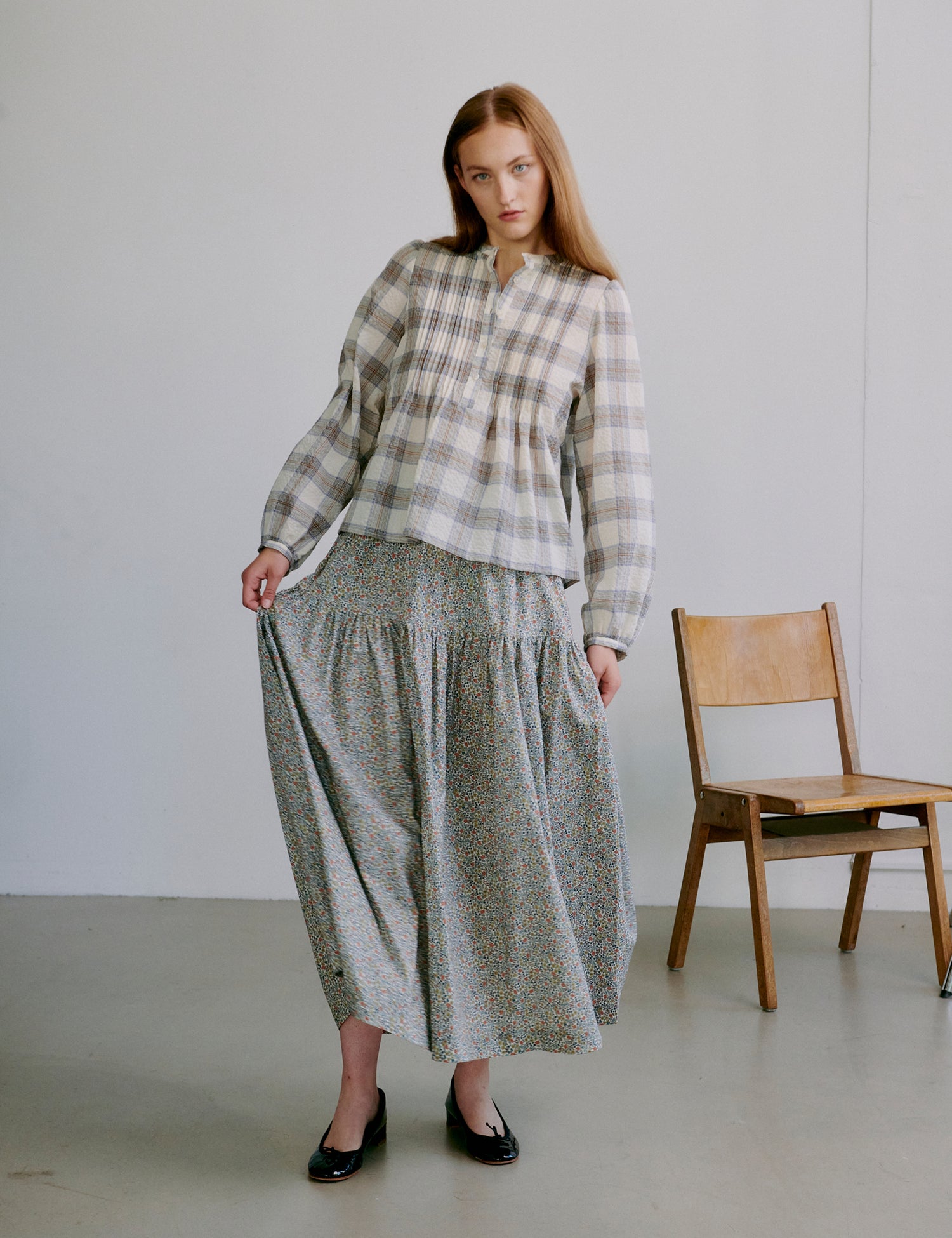 Woman wearing a check patterned white and grey shirt and long floral skirt standing in a minimalistic room with a wooden chair.