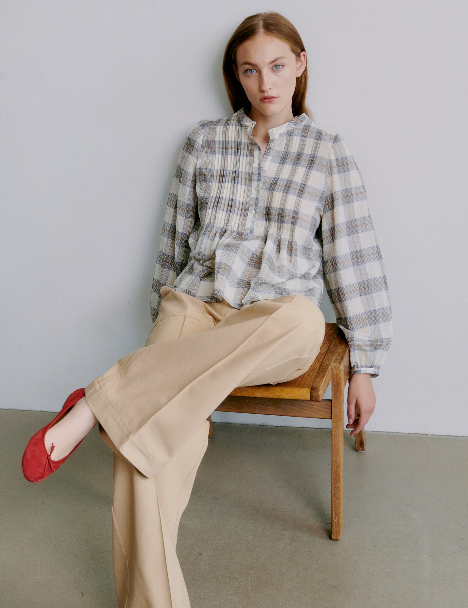 Woman sitting on a wooden chair wearing a plaid shirt and beige pants with red shoes.
