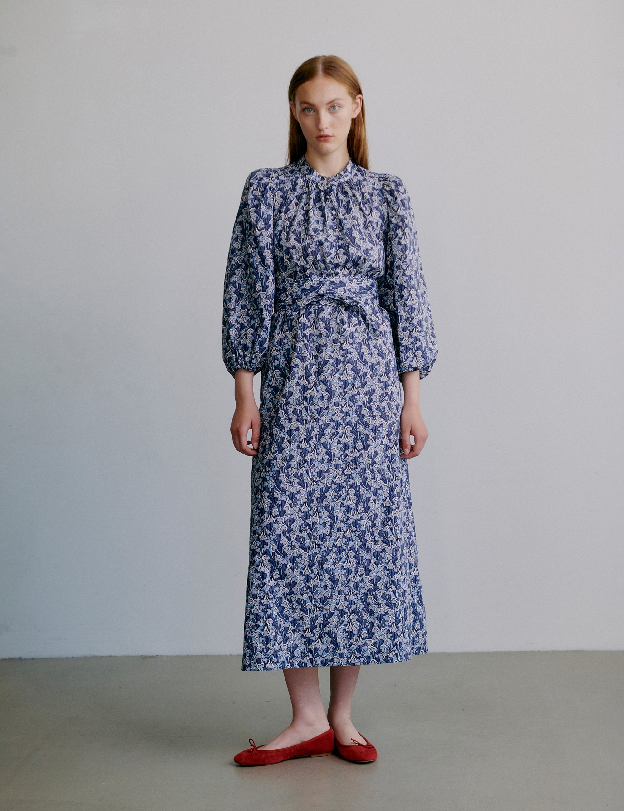 Woman wearing a blue floral dress with long sleeves, matching belt and red shoes against a plain background