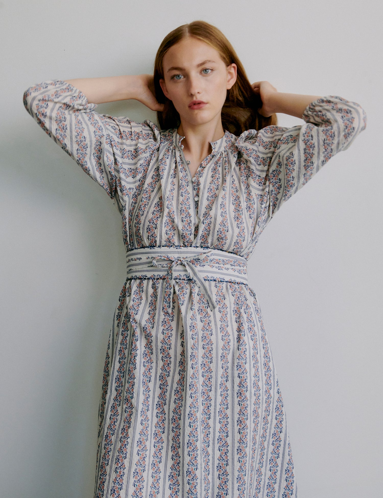 Woman wearing a floral and stripe patterned dress with long puff sleeves and matching belt against a plain background