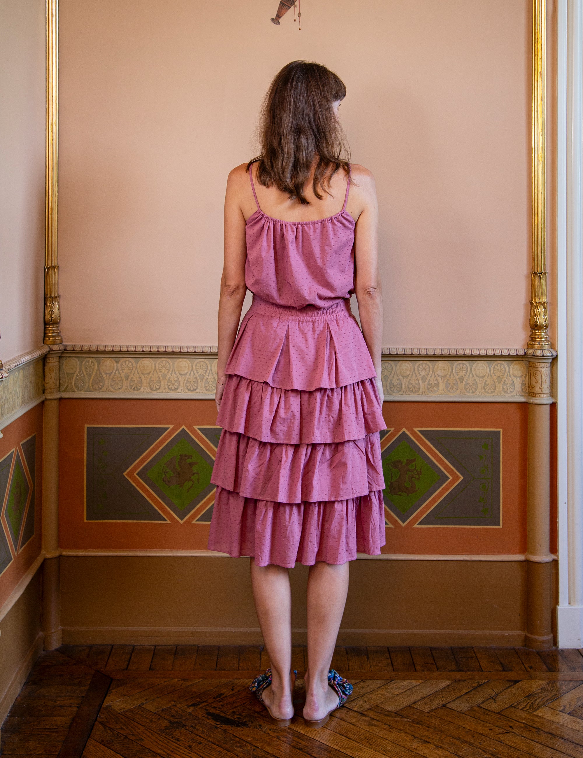 Woman wearing a pink tiered skirt and matching strap top  standing in an ornate room with decorative walls.
