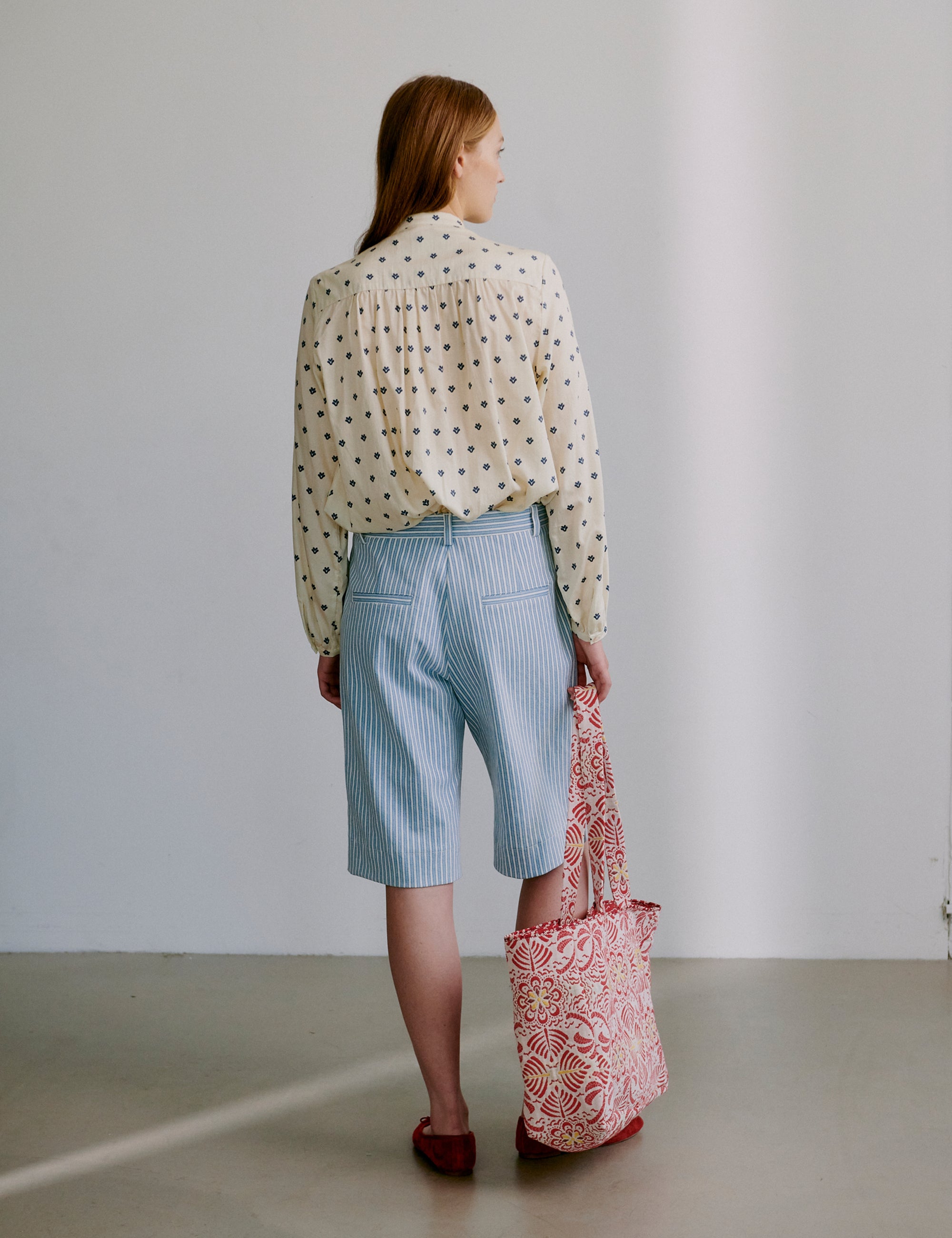 Woman in long sleeved polka dot patterned shirt and light blue and white striped shorts holding a red floral patterned tote bag 