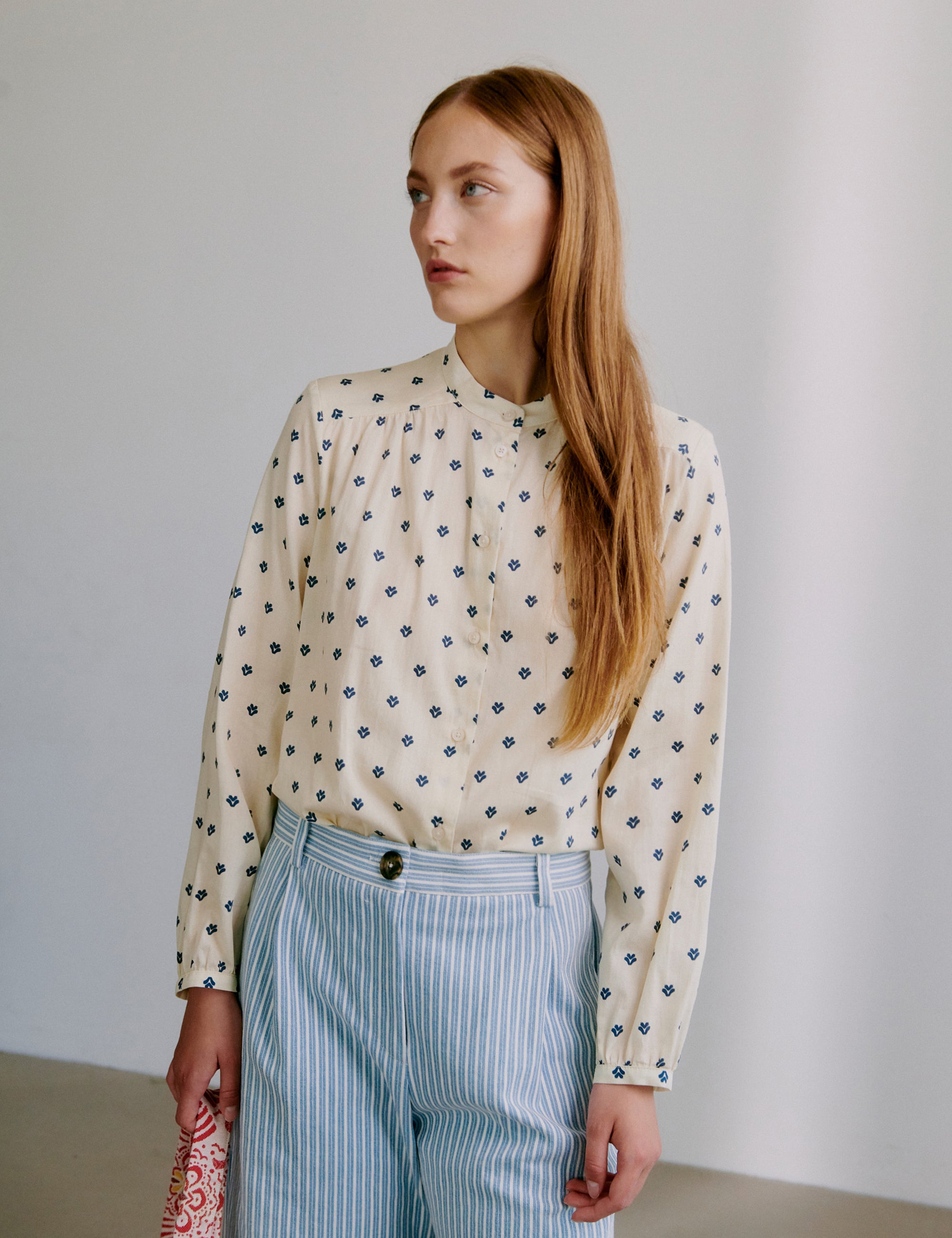 Woman wearing a beige patterned long sleeve blouse and striped pants against a plain background