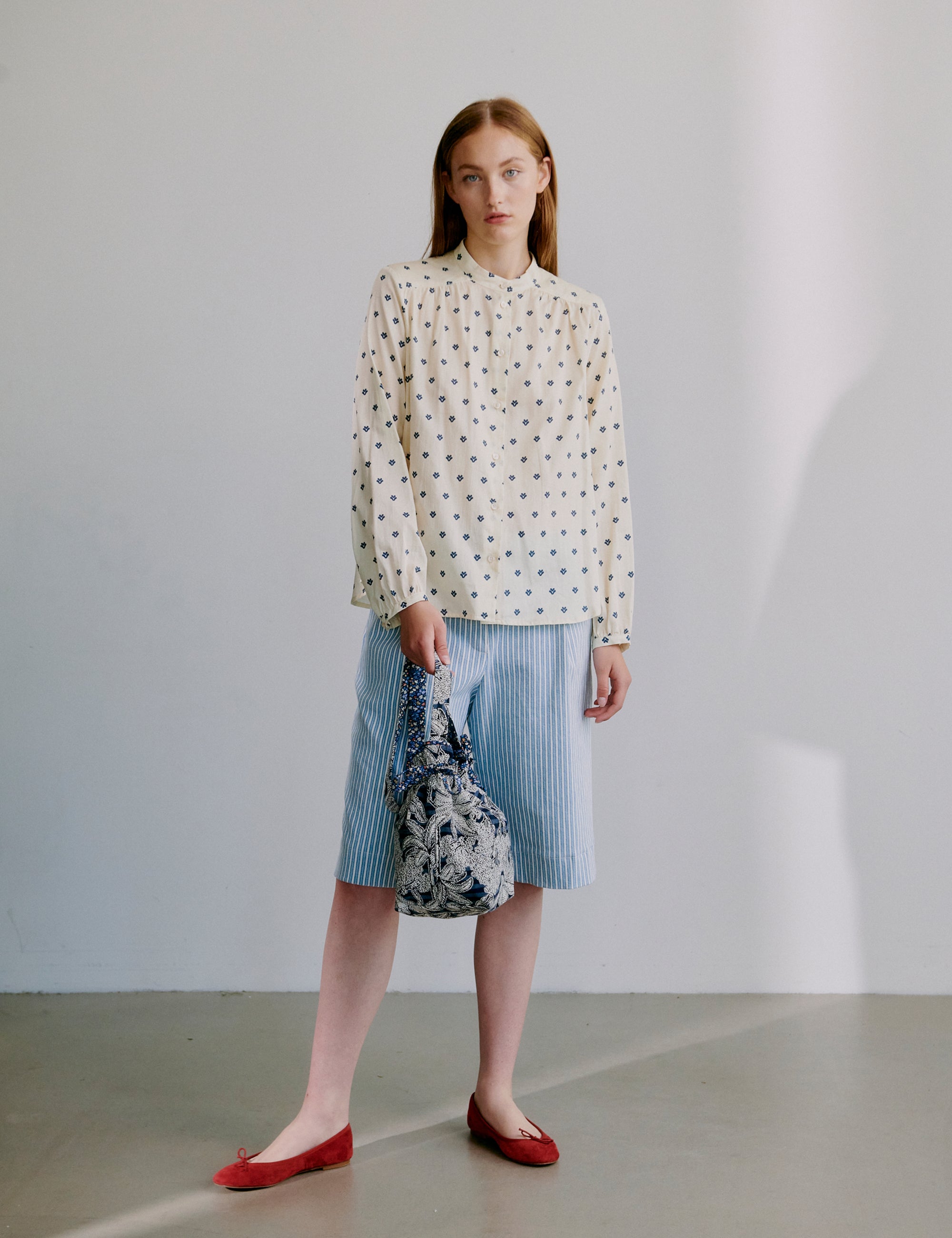 Woman wearing a beige polka dot patterned long sleeve blouse and striped skirt holding a blue floral fabric bag 