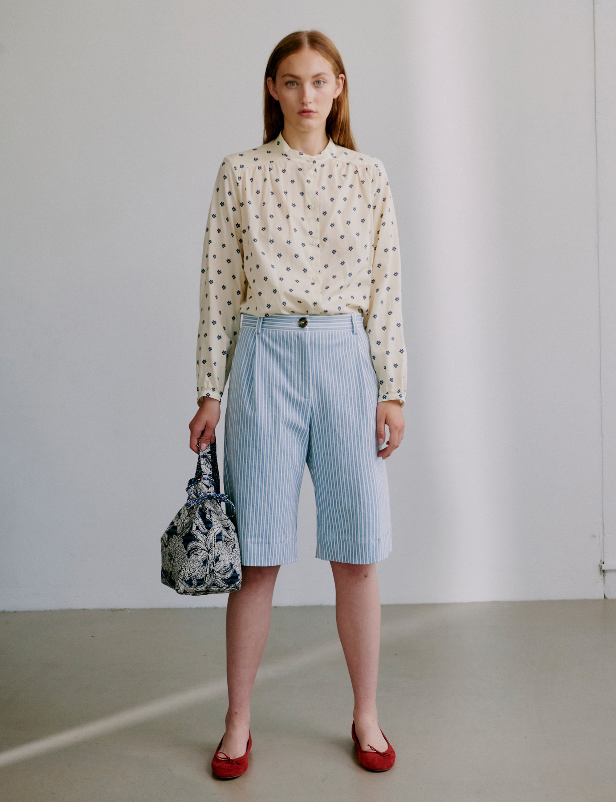 Woman wearing a polka dot patterned blouse and striped shorts, holding a floral handbag against a plain background