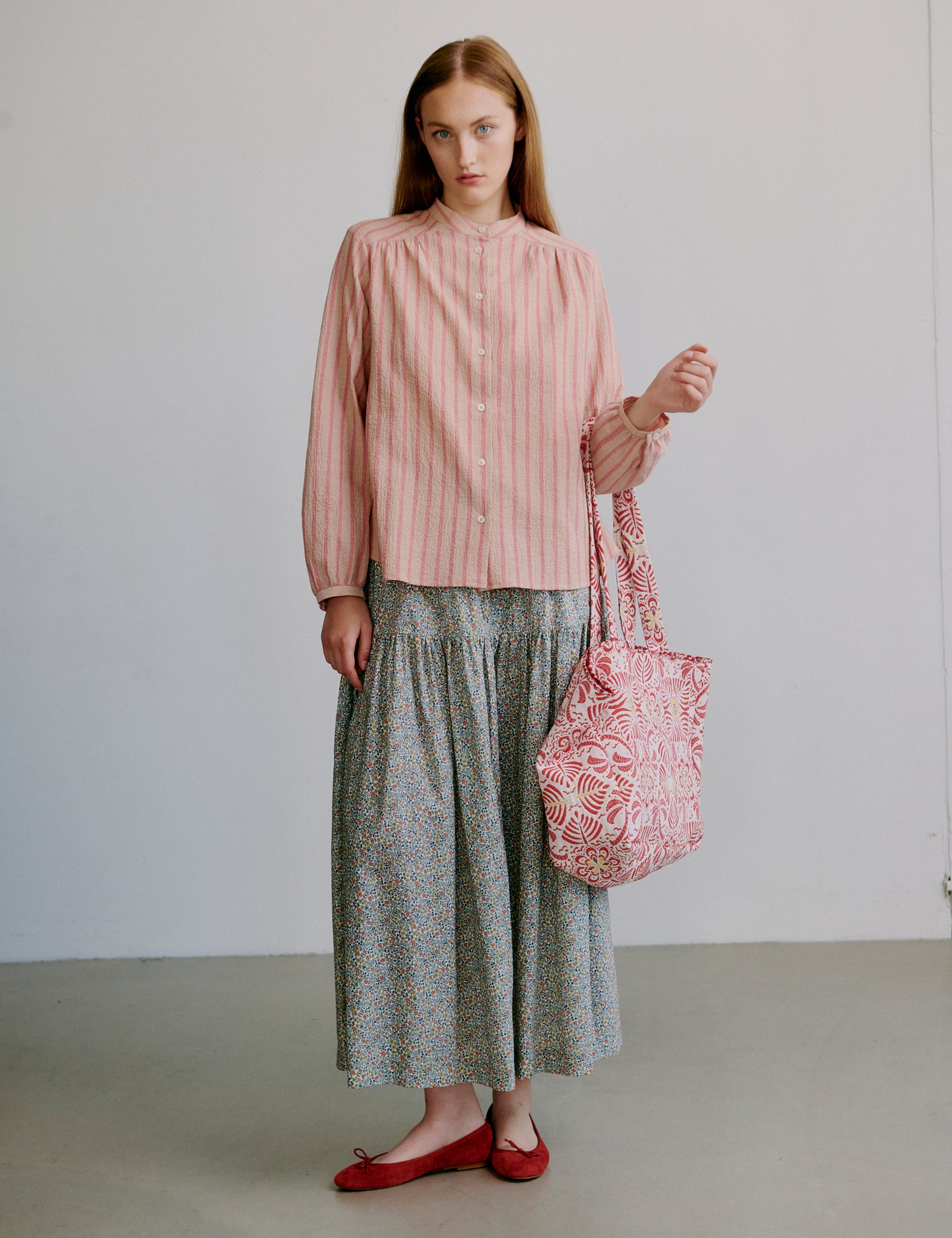 Woman wearing a pink striped long sleeve shirt and flower patterned skirt holding a red patterned tote bag