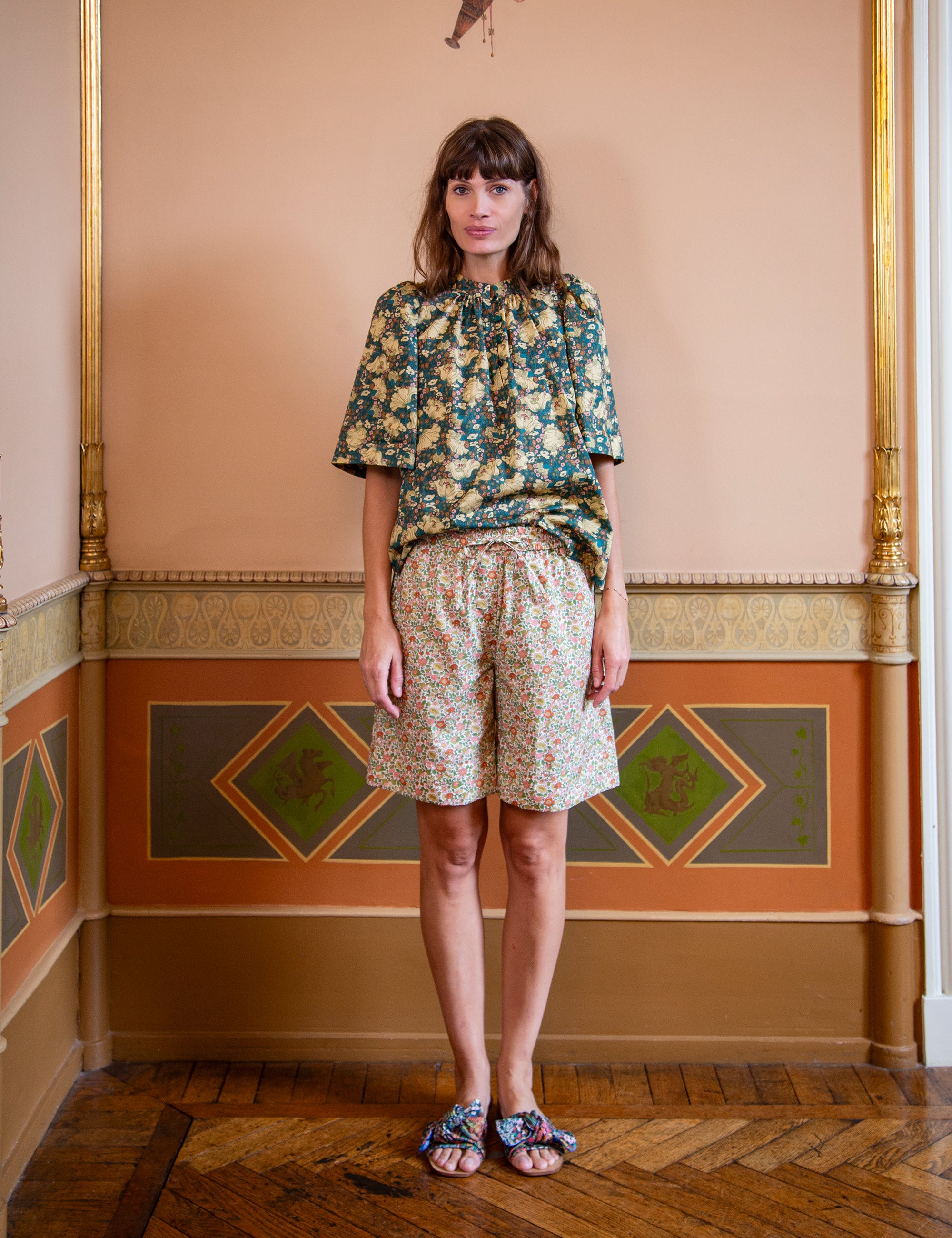 Woman wearing a floral top and shorts  standing in a room with decorative walls.