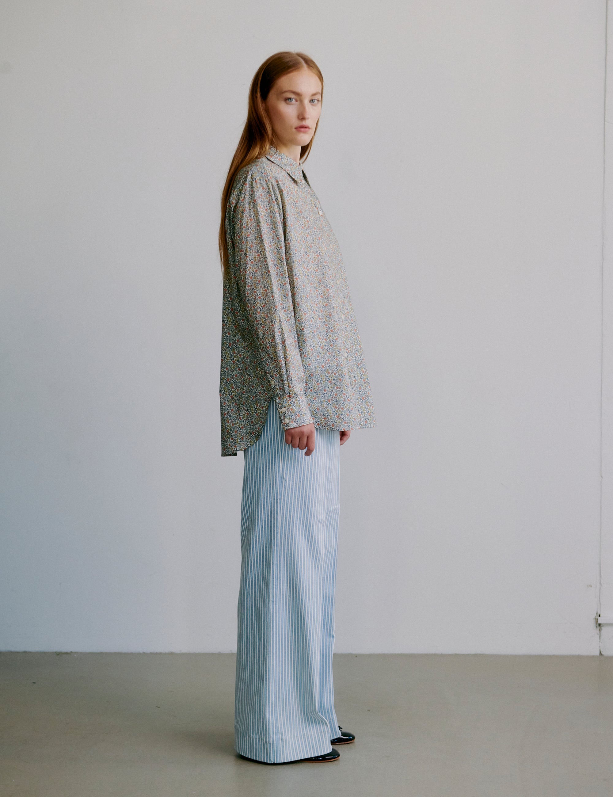 Woman wearing a floral patterned long sleeve shirt and light blue striped pants against a plain background
