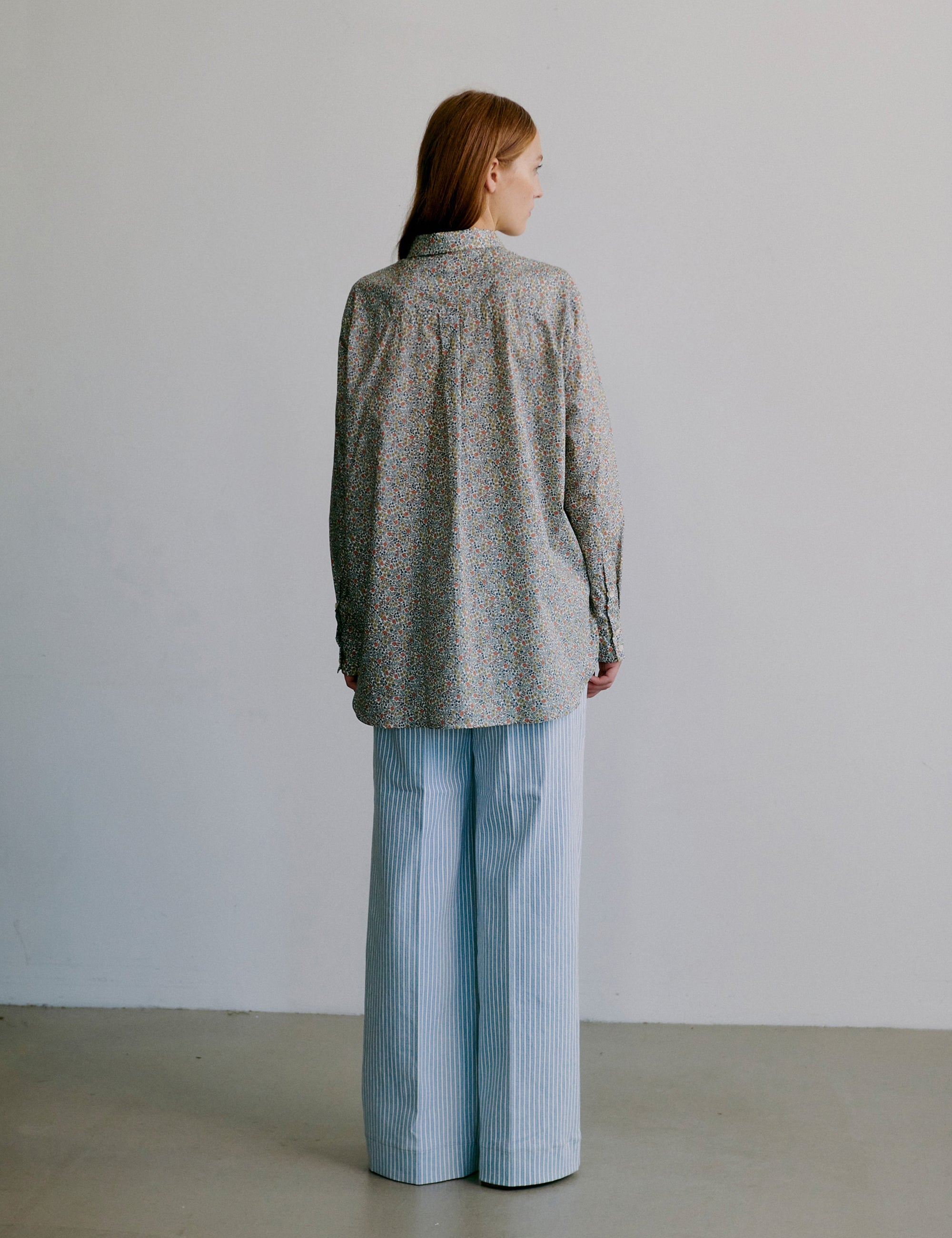 Woman wearing a floral patterned long sleeve shirt and light blue striped pants against a plain background