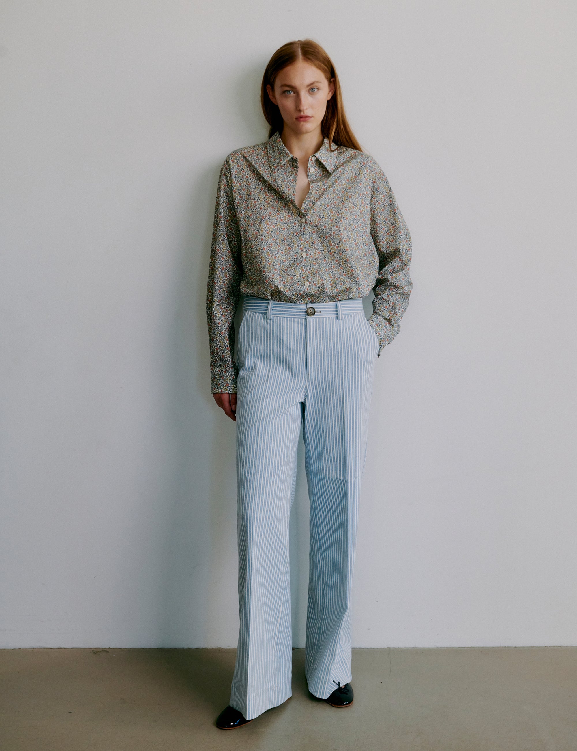 Woman wearing a floral patterned long sleeve shirt and light blue striped pants against a plain background