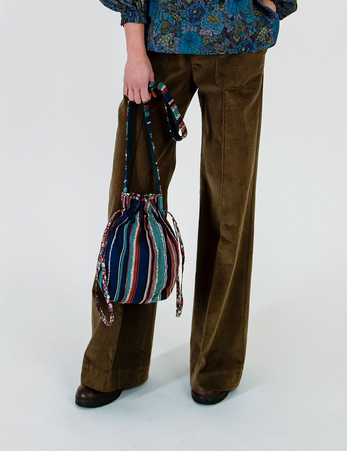 Woman wearing brown corduroy trousers and a blue floral top holds a small striped bucket bag with a drawstring closure against a plain light gray studio background.