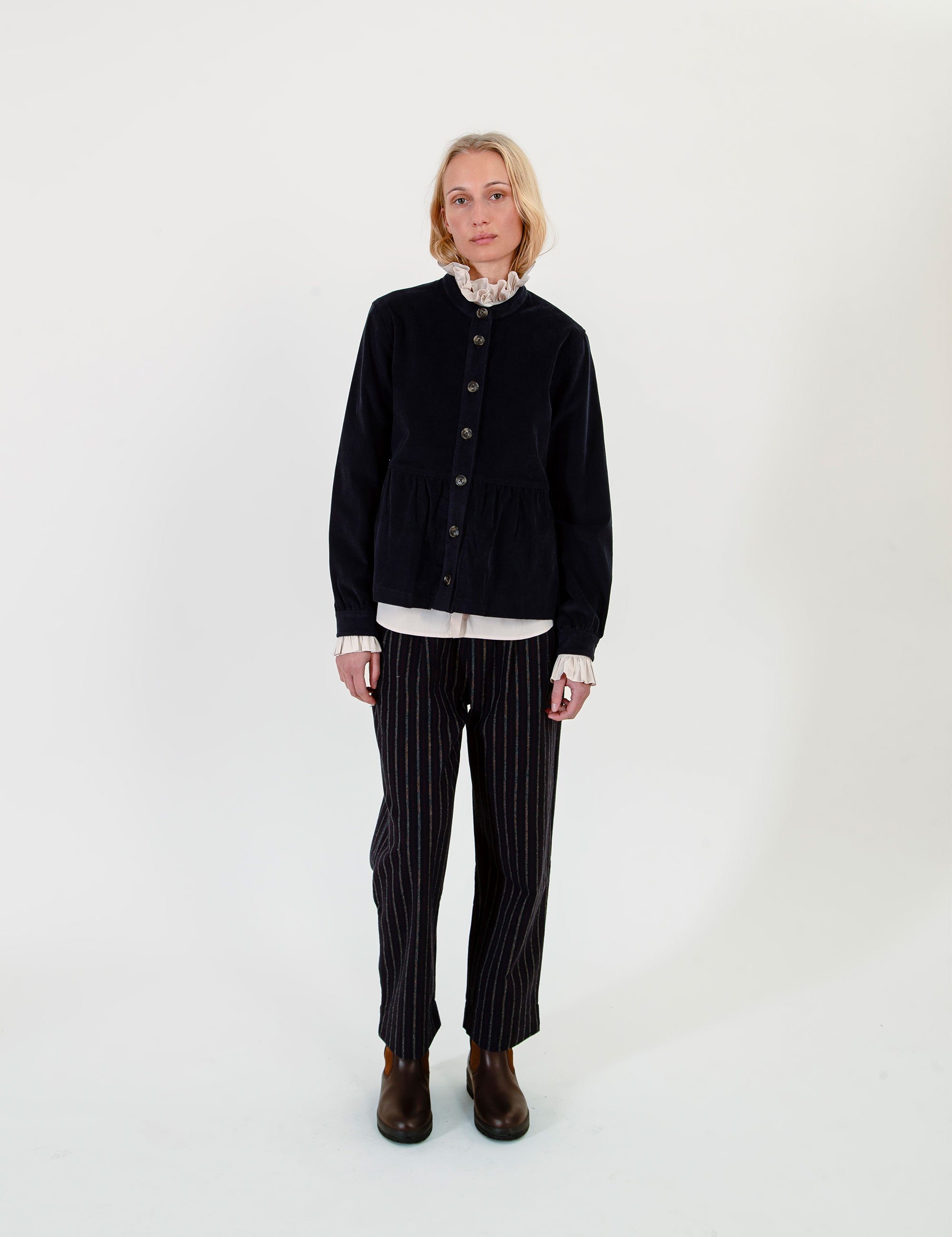 A woman wearing a dark navy corduroy jacket over a white ruffled shirt, paired with matching navy pinstripe trousers and brown boots.