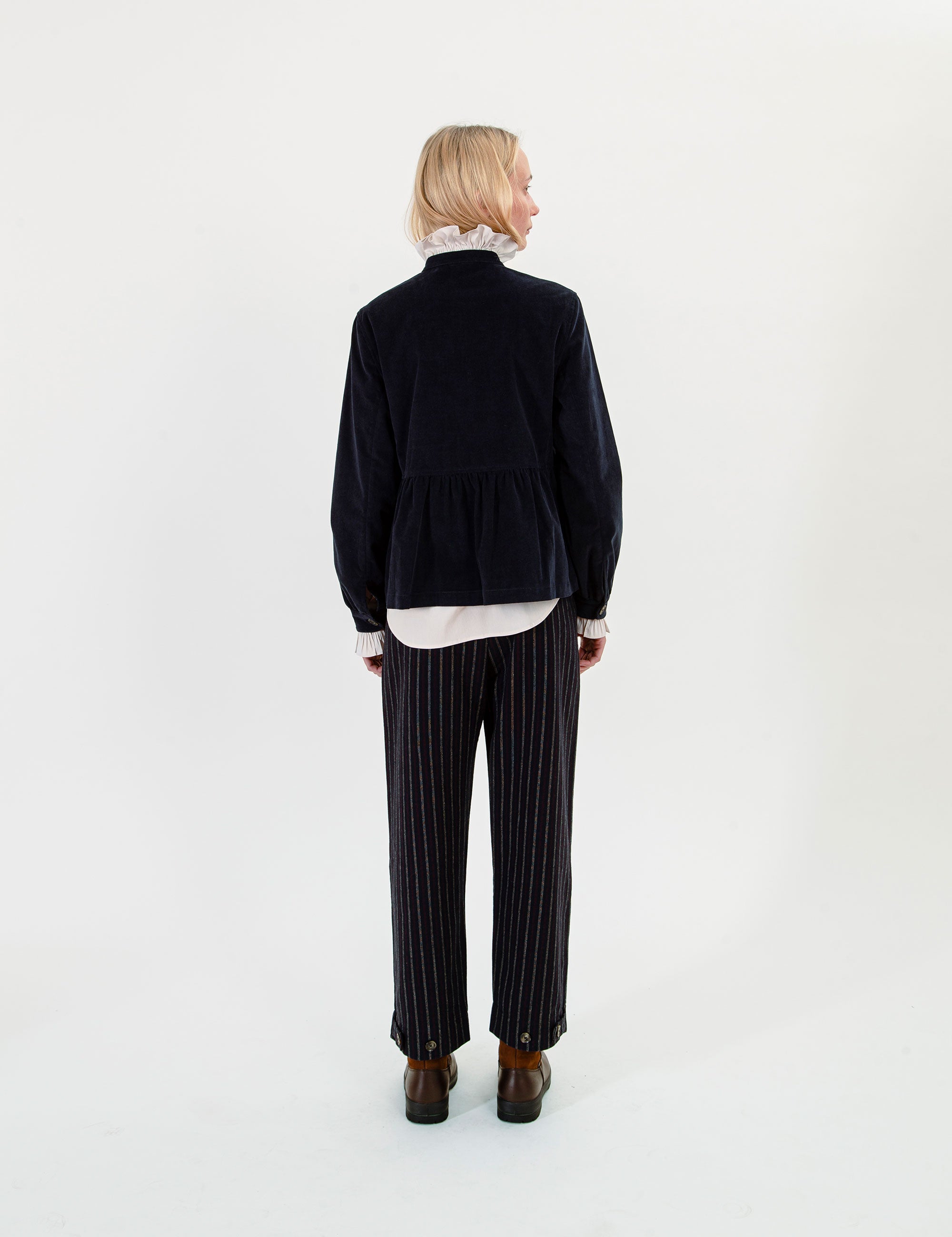 A woman wearing a dark navy corduroy jacket over a white ruffled shirt, paired with matching navy pinstripe trousers and brown boots.