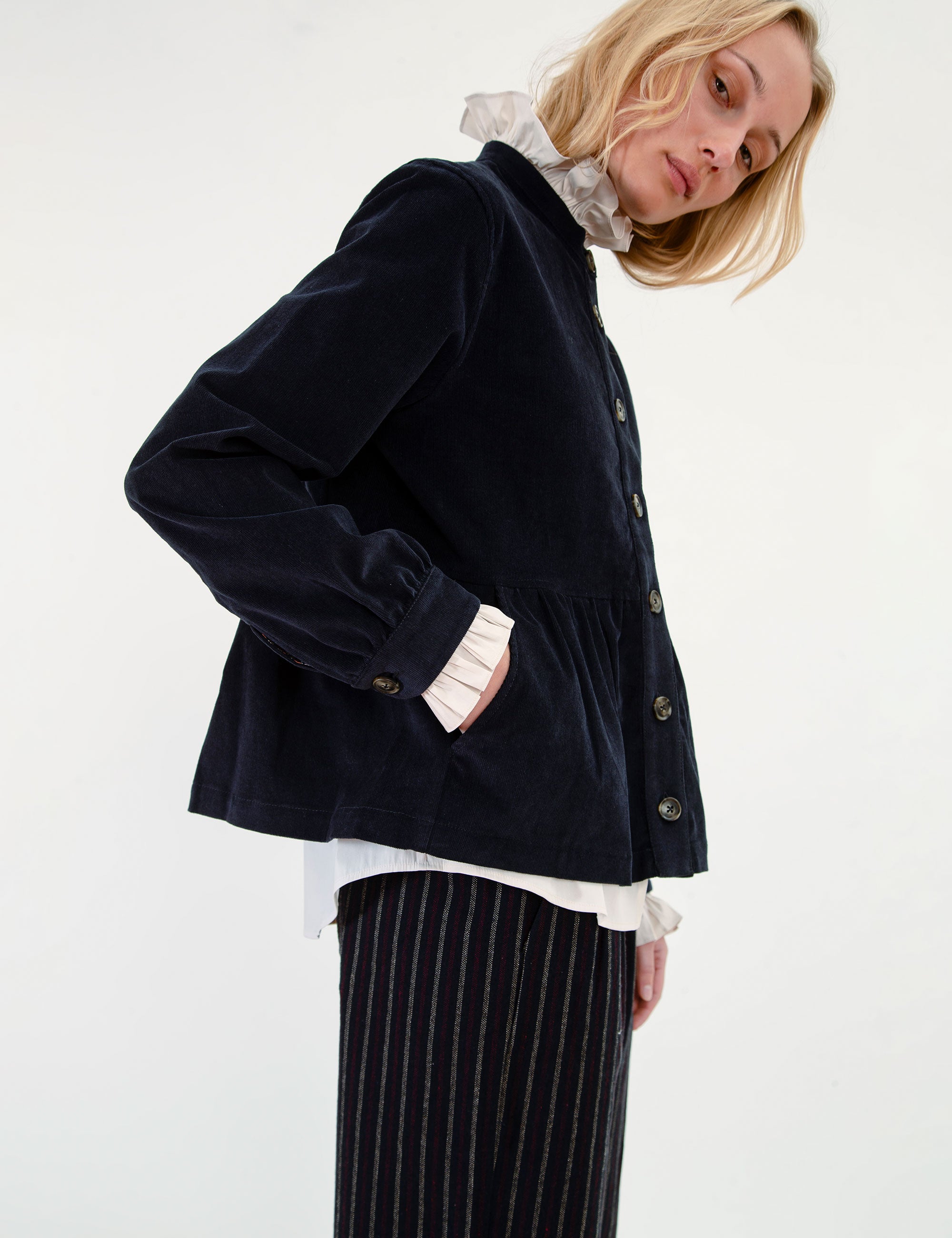A woman wearing a dark navy corduroy jacket over a white ruffled shirt, paired with matching navy pinstripe trousers and brown boots.