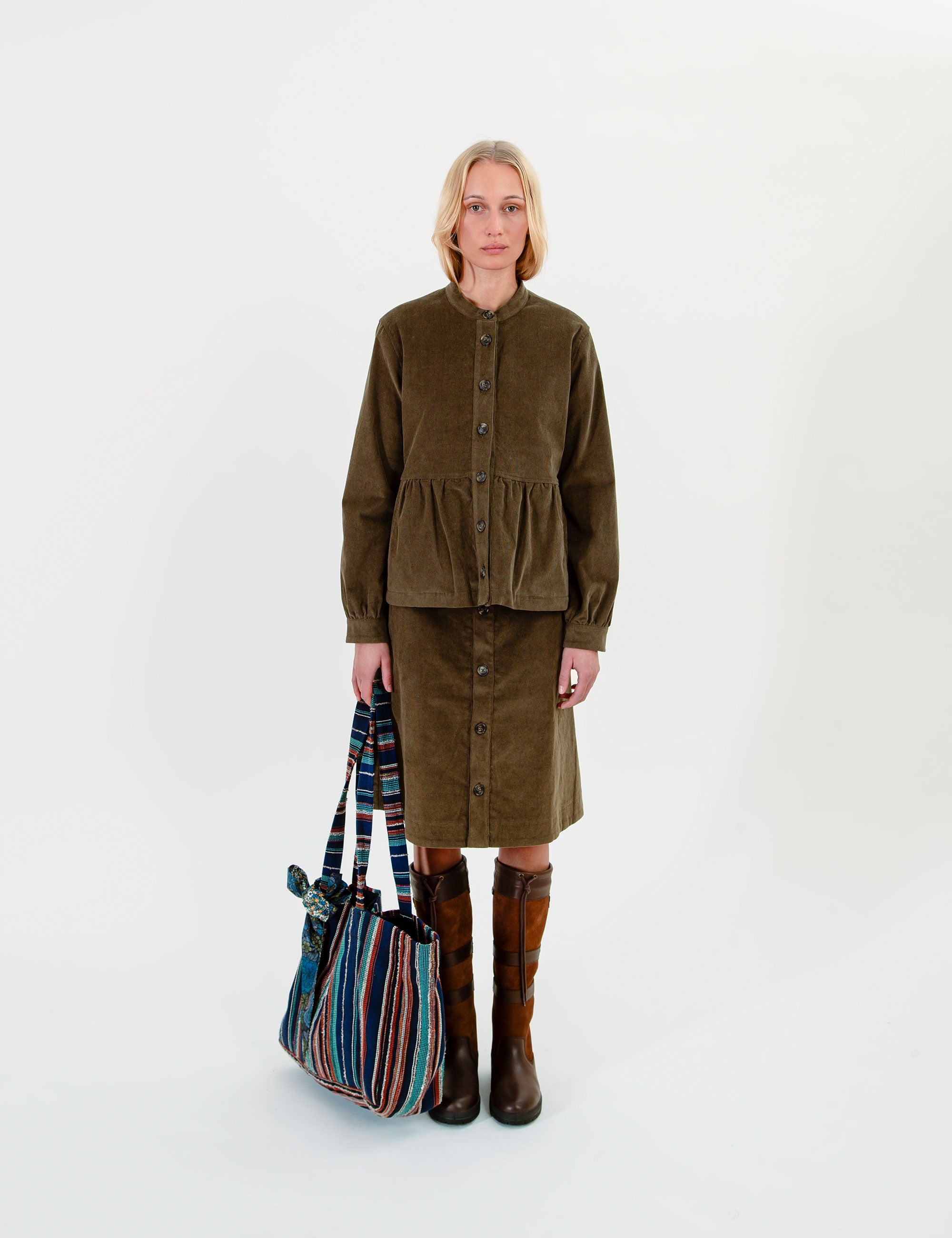 A woman wearing an olive-green corduroy jacket and matching skirt set, brown knee-high boots, and holding a striped tote bag.