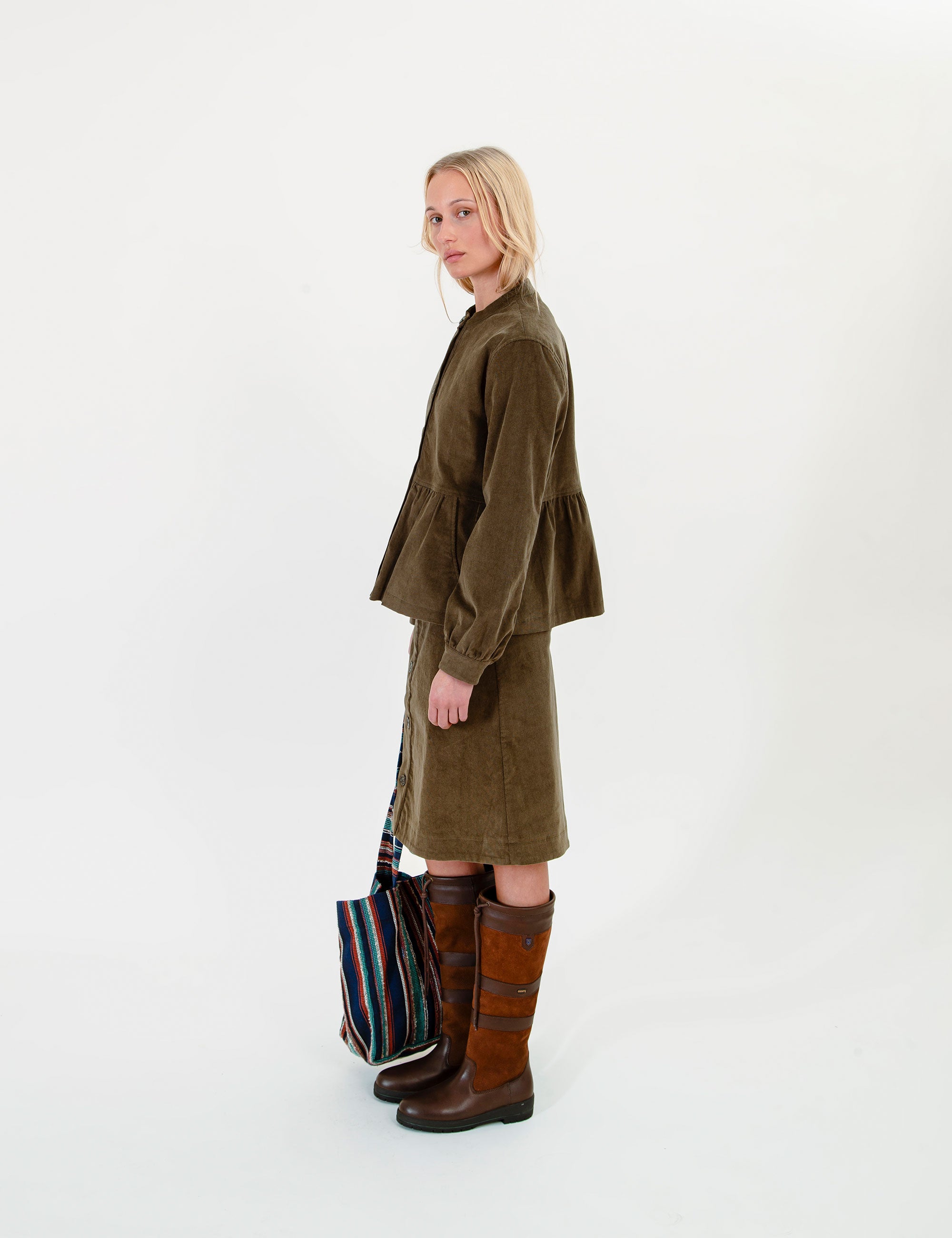 A woman wearing an olive-green corduroy jacket and matching skirt set, brown knee-high boots, and holding a striped tote bag.