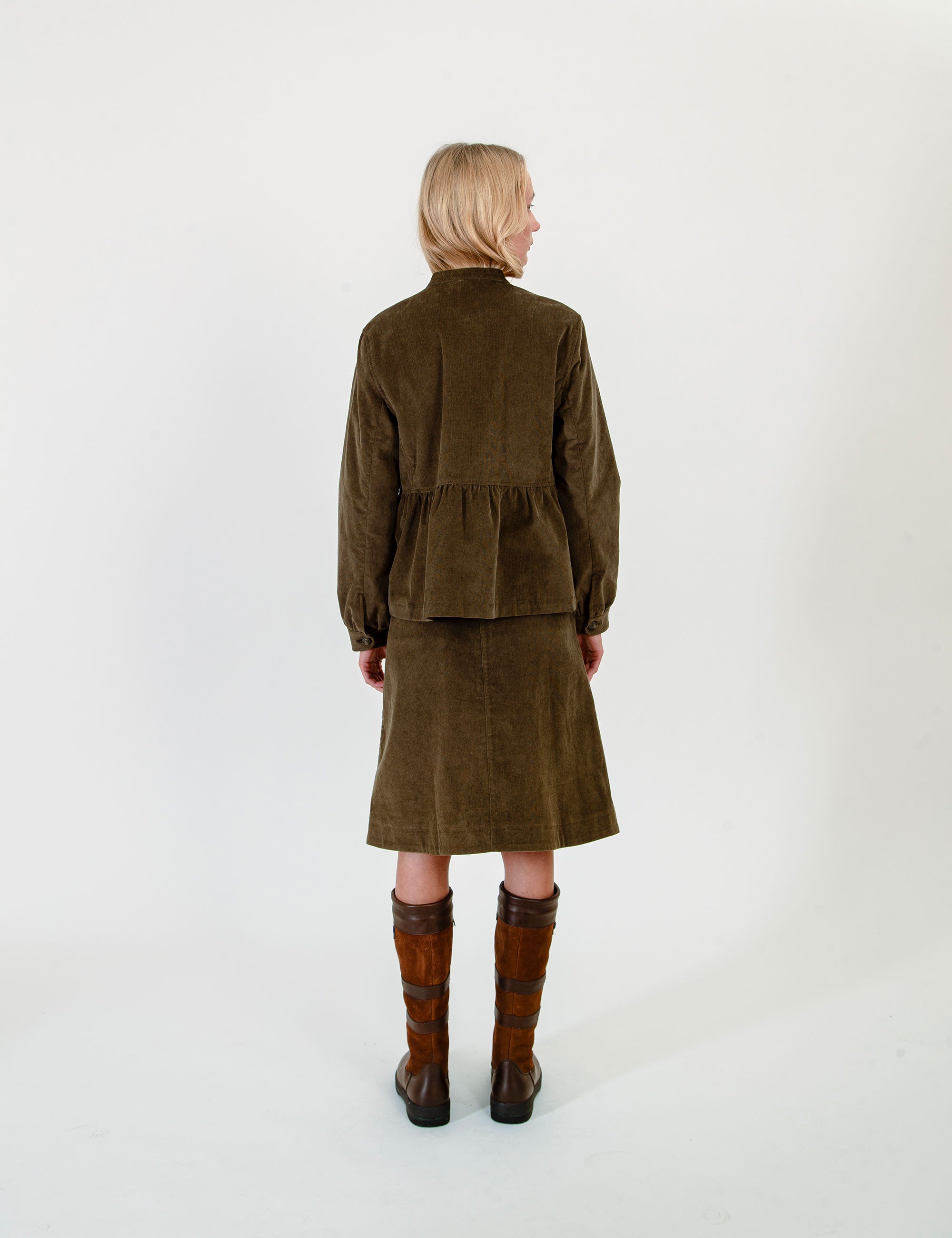 A model viewed from the back wearing a green corduroy jacket and matching skirt set, paired with brown leather knee-high boots, against a plain white studio background.