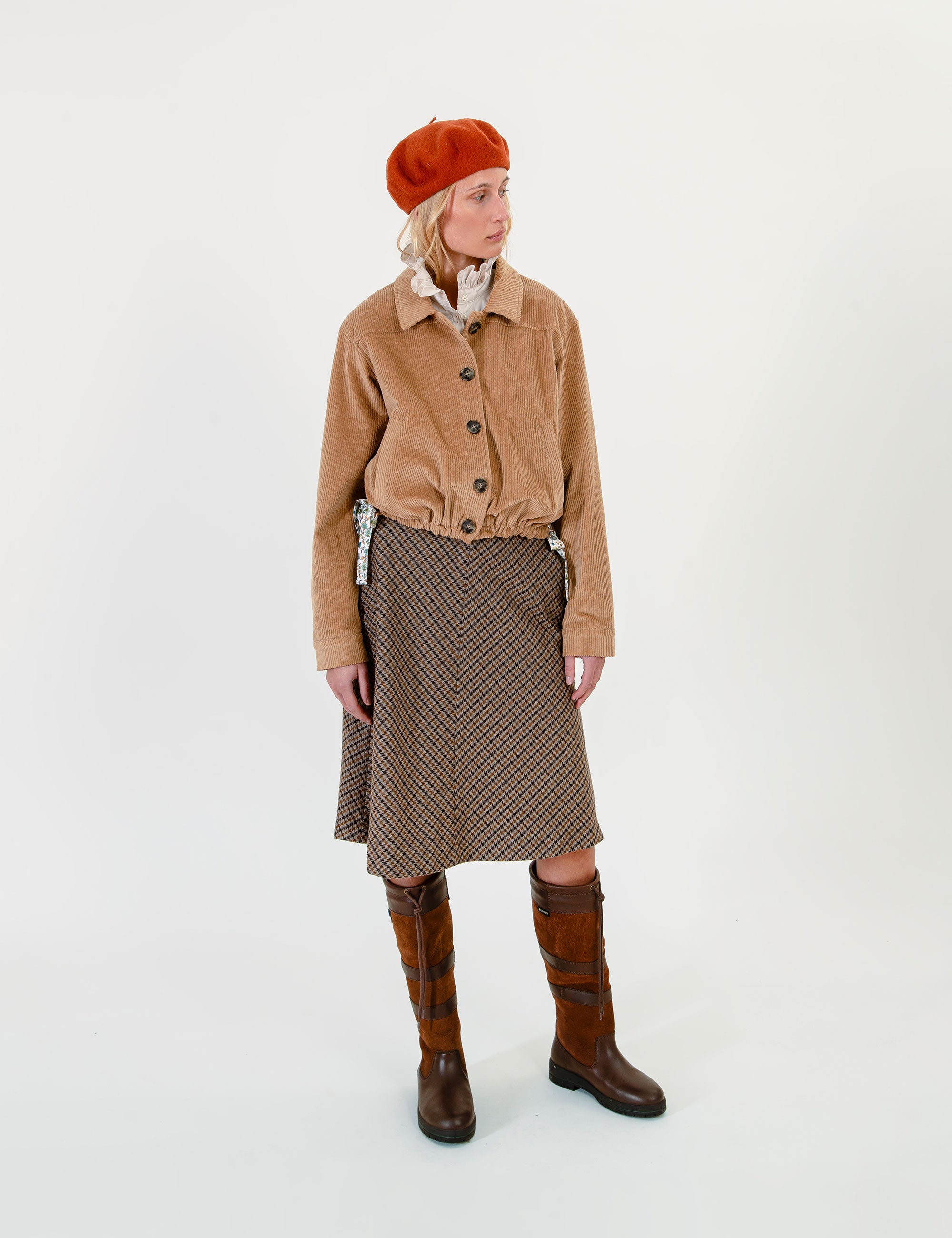 A woman wearing a tan corduroy jacket, a brown patterned skirt, brown leather boots, and an orange-red beret against a plain white studio background.