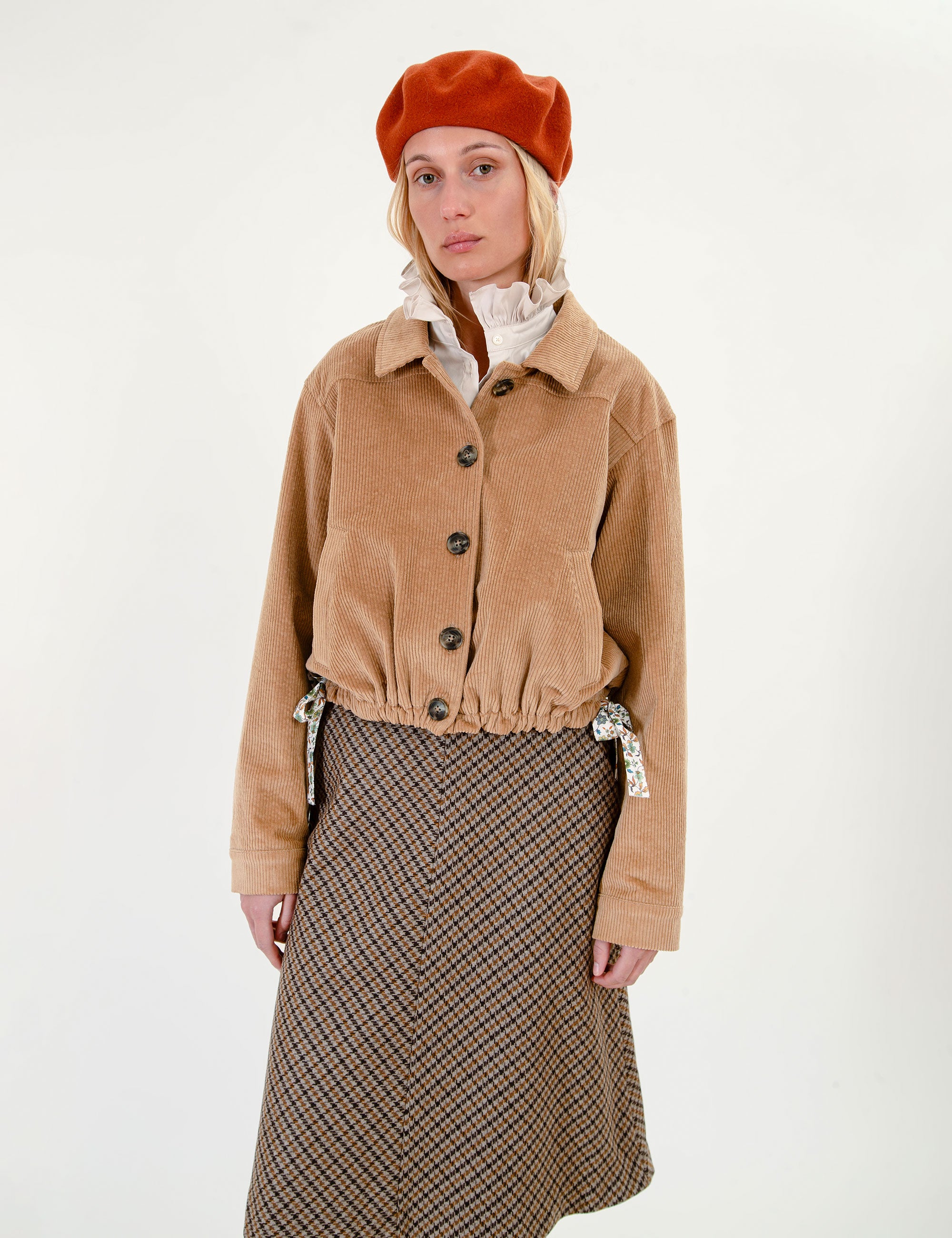 Woman wearing a tan jacket and plaid skirt with an orange beret on a white background