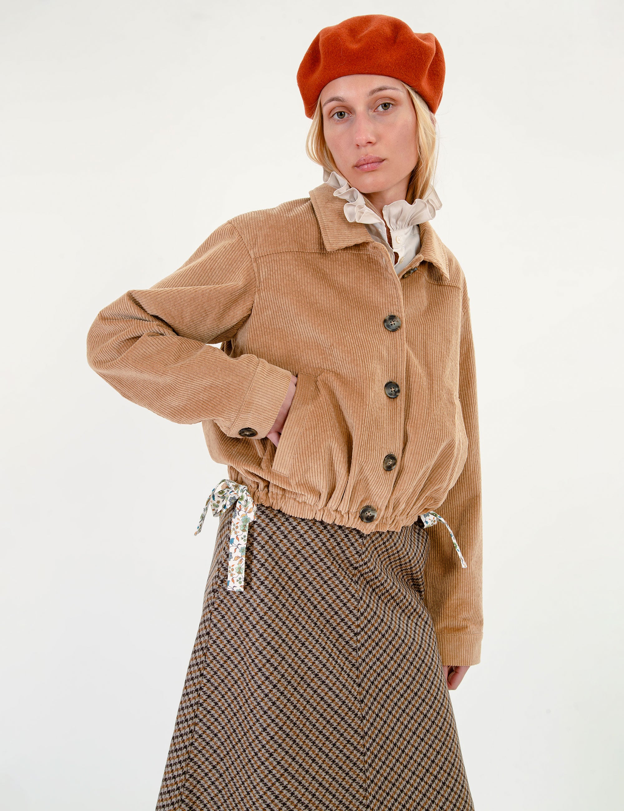 Woman wearing a cropped tan corduroy jacket with dark buttons, a brown and beige checkered skirt, a cream ruffled collar, and an orange-red beret, photographed against a plain white studio background.