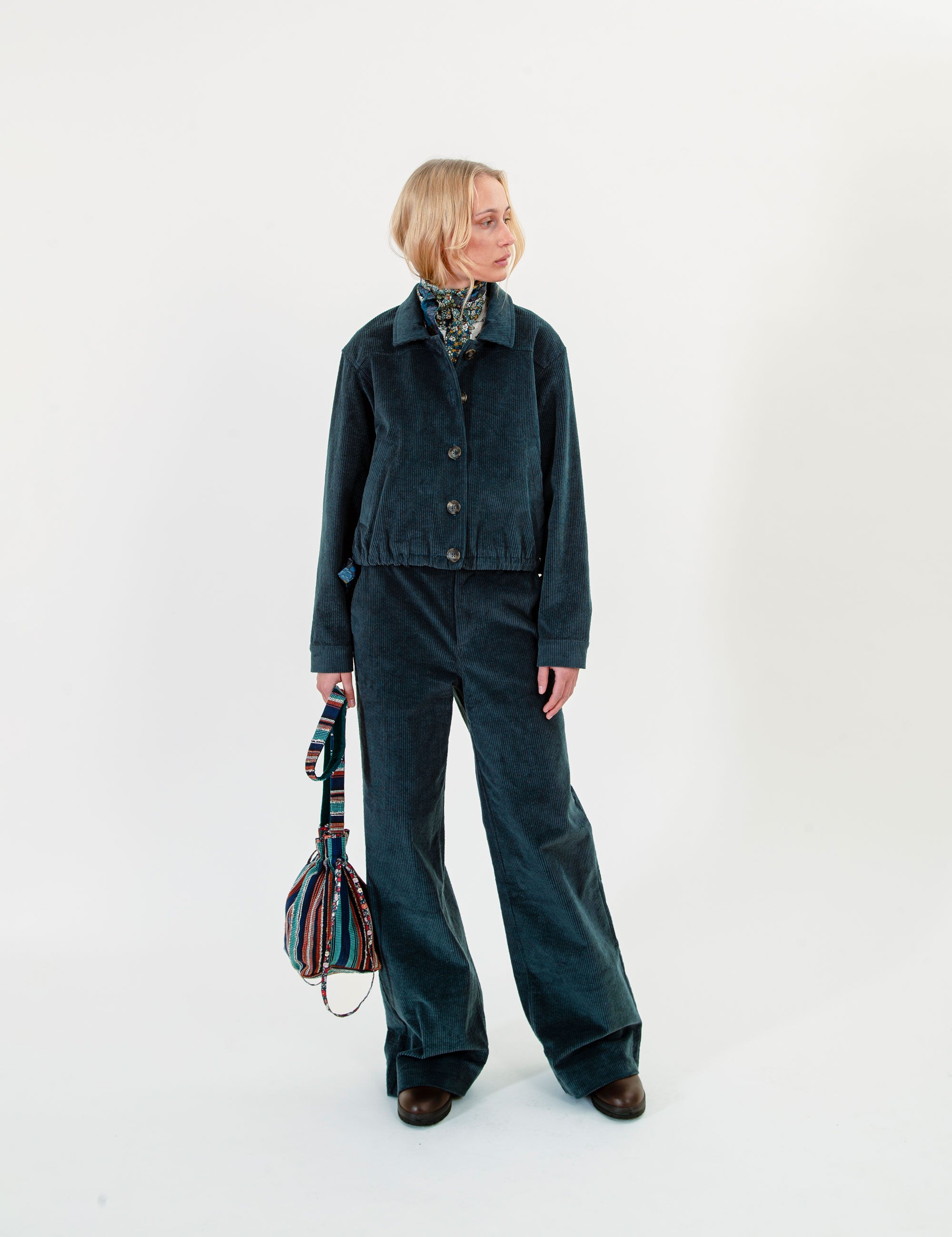 A woman stands against a plain white studio background wearing a dark teal corduroy jacket and matching wide-leg trousers, styled with a patterned scarf and holding a striped handbag.