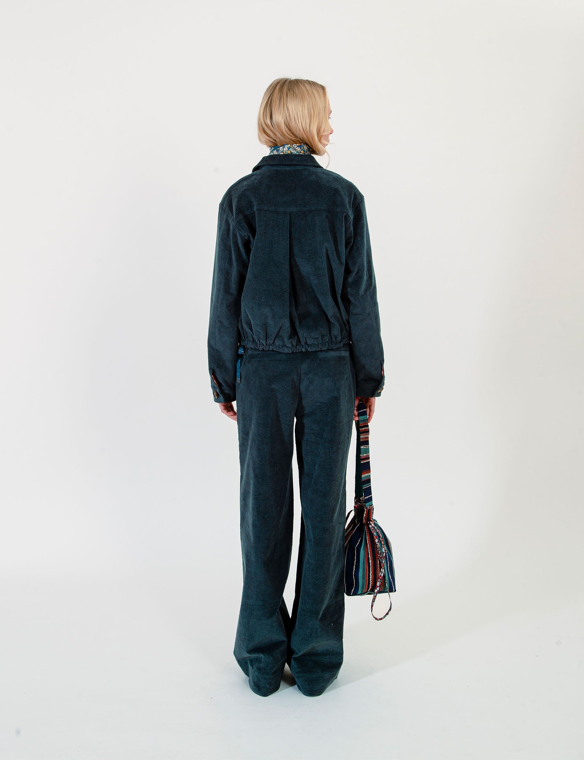 A model is shown from the back wearing a dark teal corduroy jacket and pants and holding a striped multicolor shoulder bag against a plain white studio background.