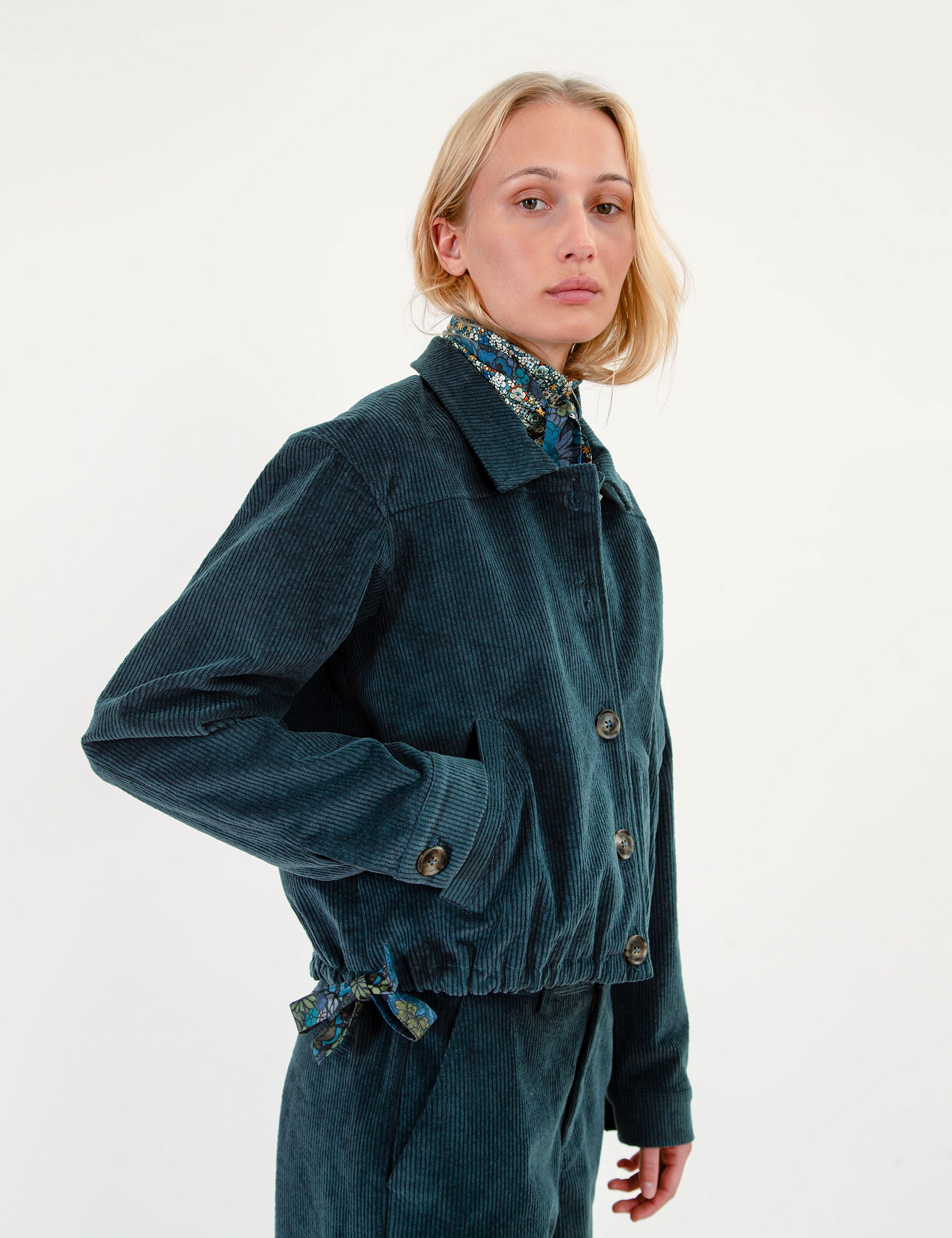 A woman wearing a dark teal corduroy jacket with a matching drawstring waist and a patterned shirt collar, photographed against a plain white studio background.