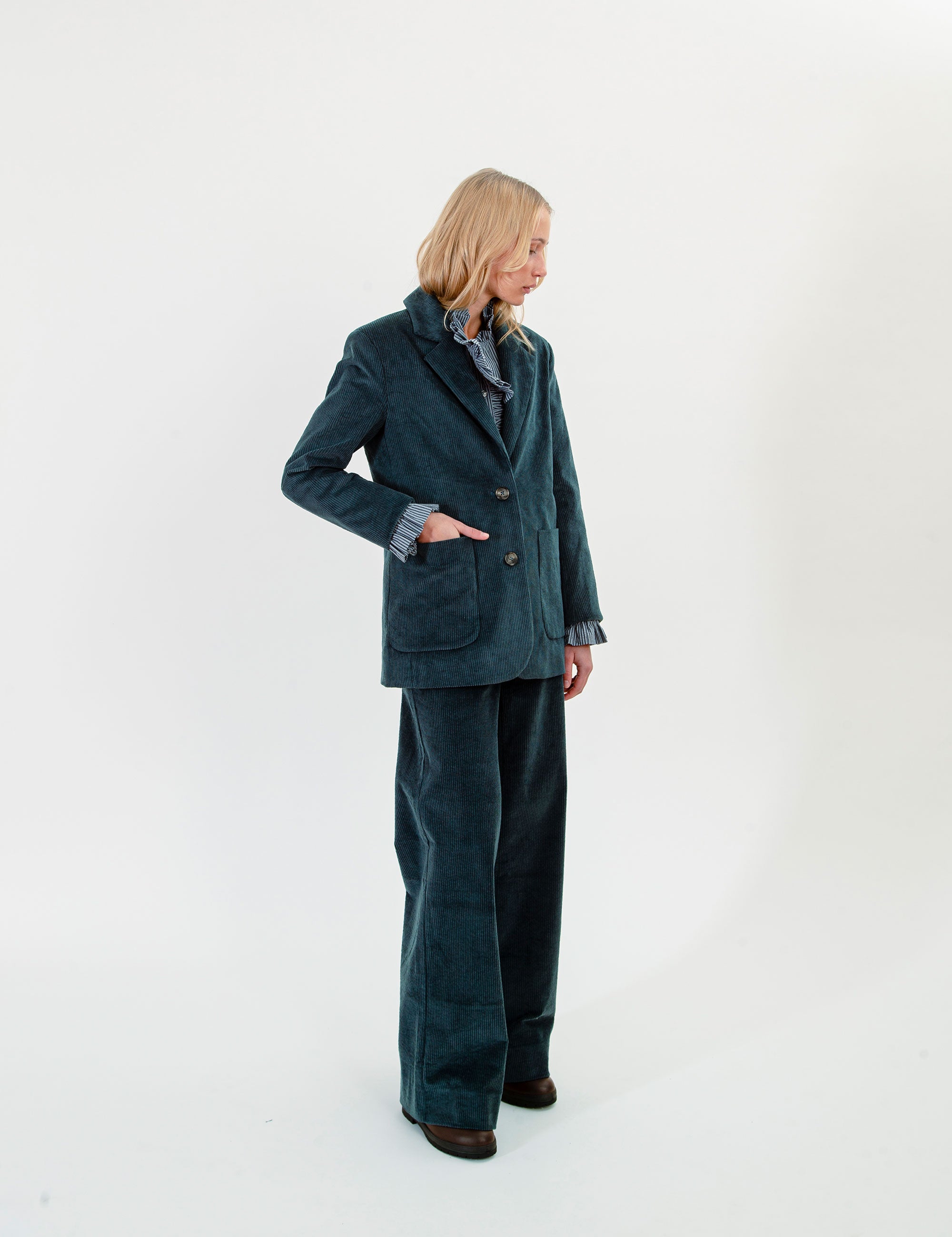 A woman stands in profile wearing a dark teal corduroy blazer and matching wide-leg trousers over a patterned collared shirt, photographed against a plain white studio background.