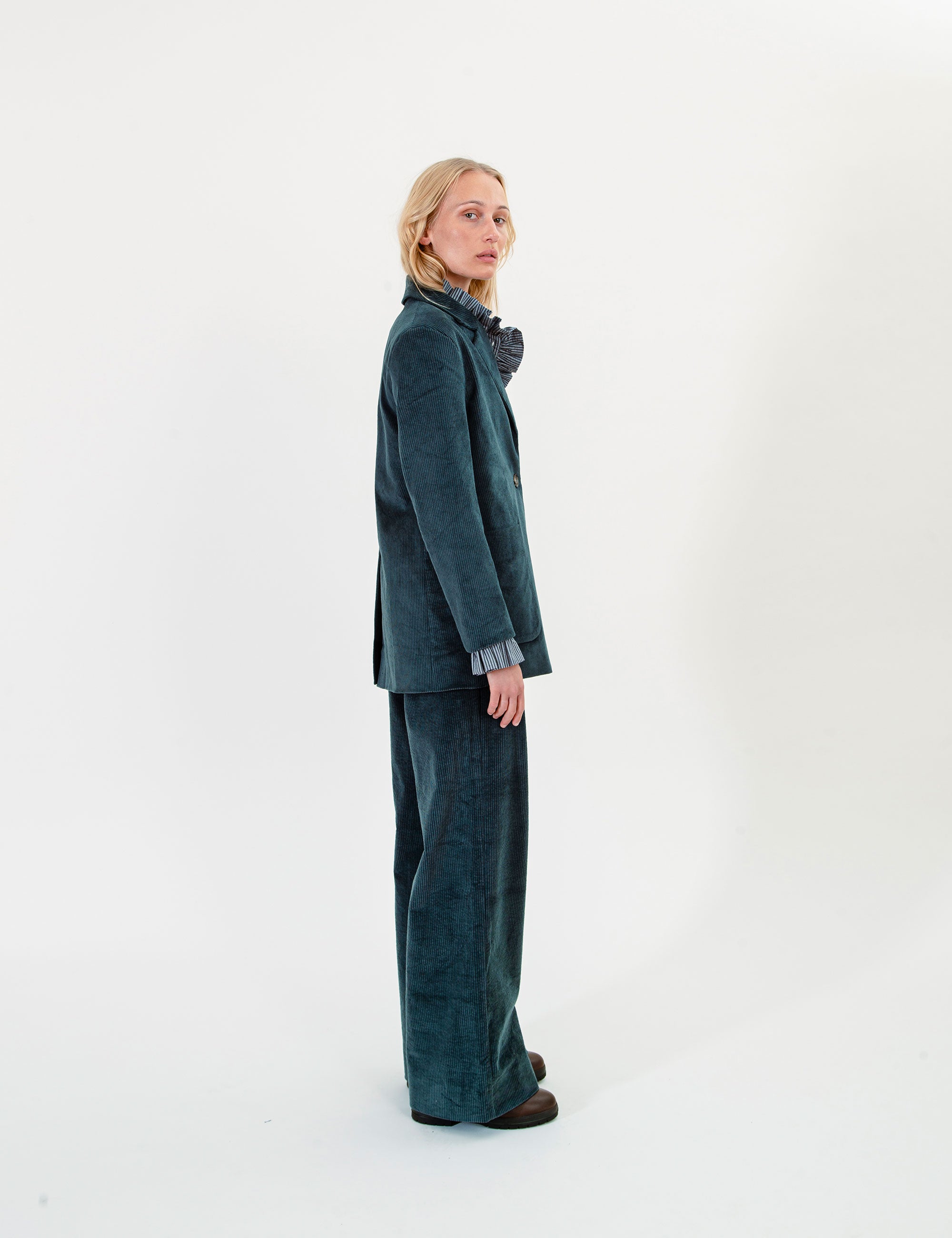 A woman stands in profile wearing a dark teal corduroy blazer and matching wide-leg trousers against a plain white studio background.