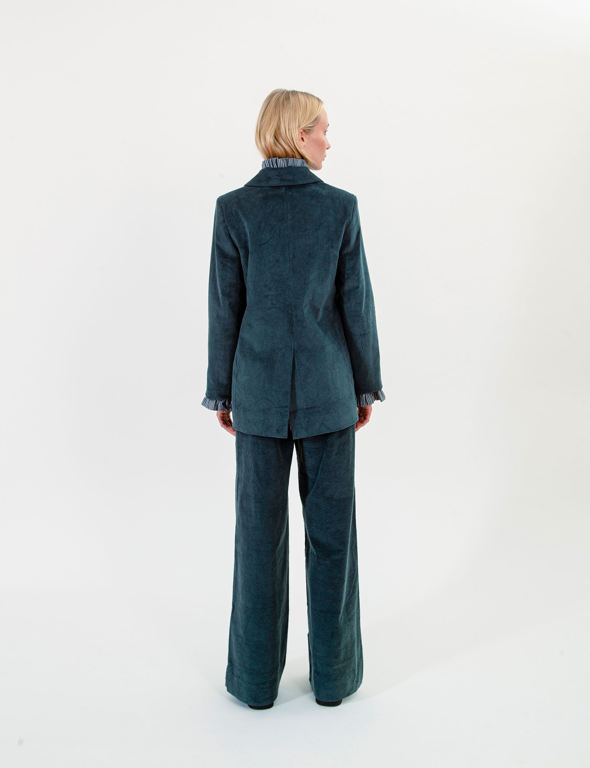 A model viewed from the back wearing a dark teal corduroy blazer and matching wide-leg trousers against a plain white studio background.