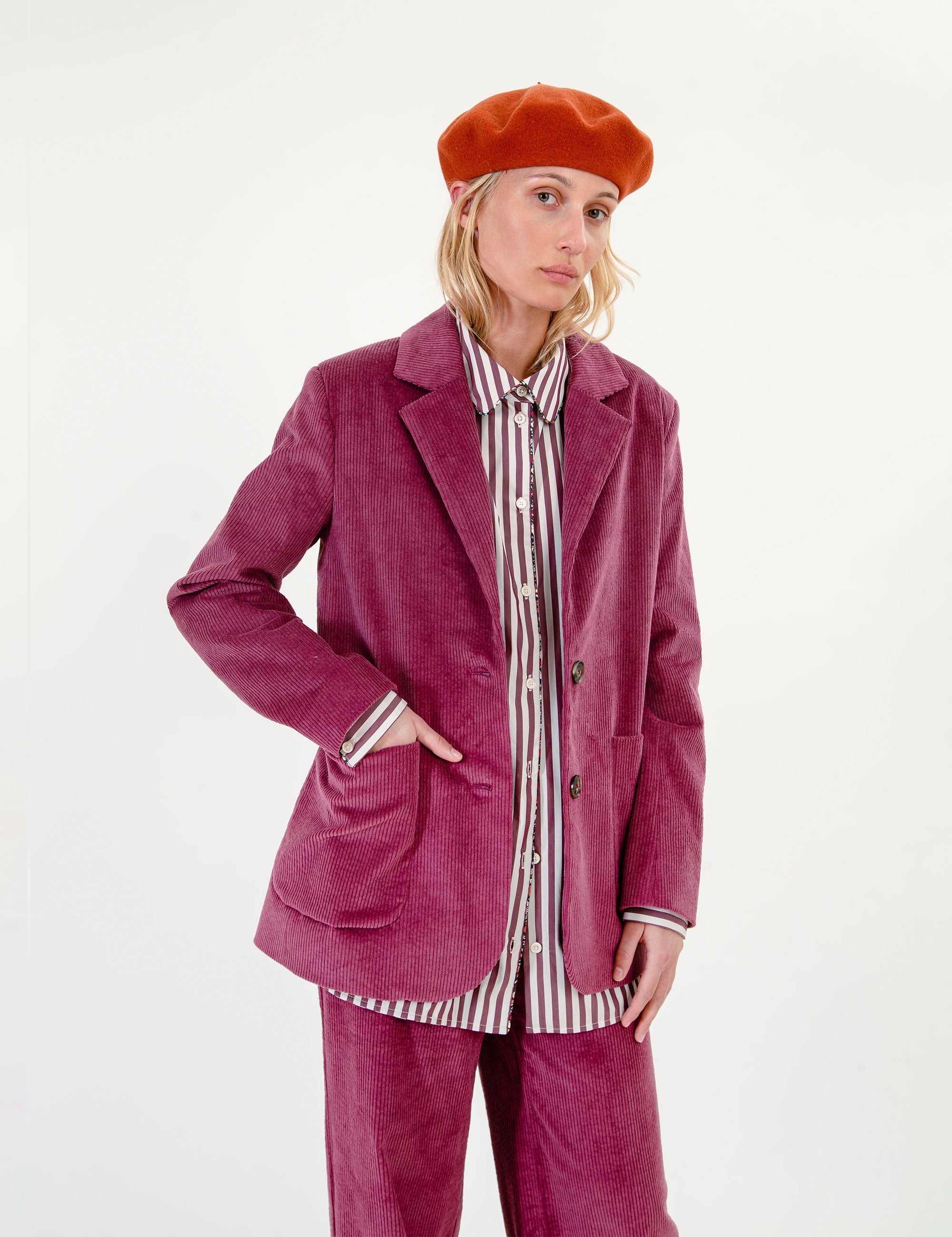 A woman wearing a matching magenta corduroy blazer and trousers set, styled with a striped shirt and a rust-orange beret, against a plain white studio background.